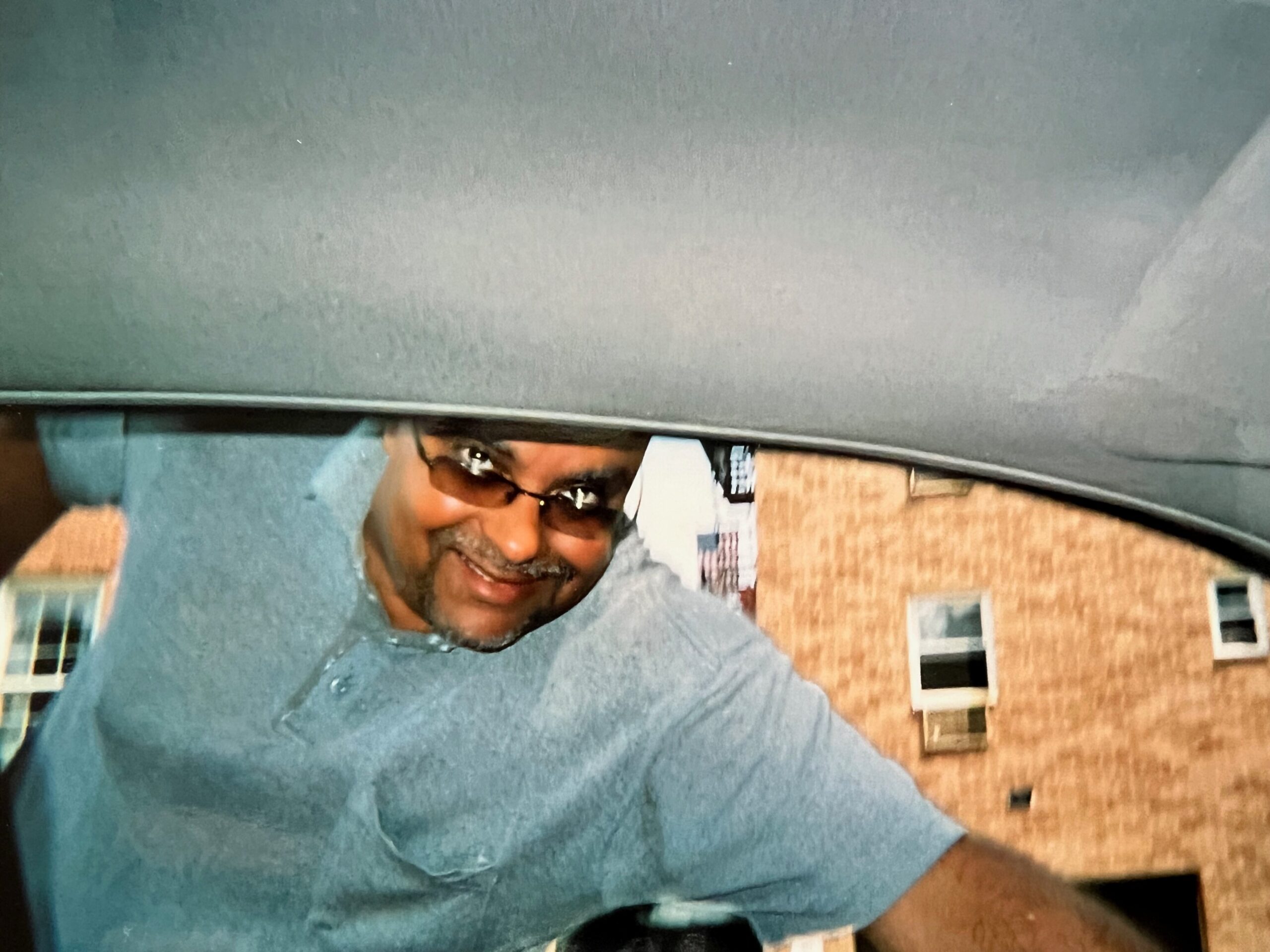 Smiling man wearing sunglasses and a gray shirt outdoors with a brick building behind him.