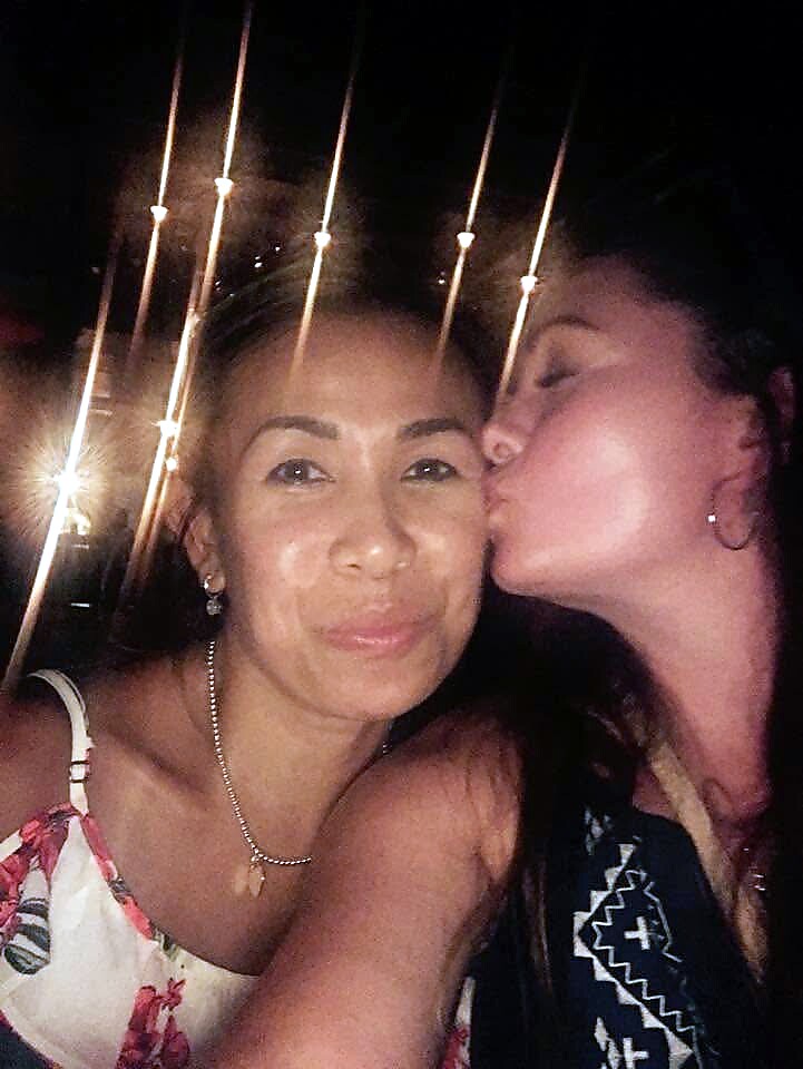 Two women posing at night; one kisses the other on the cheek against a backdrop of string lights.