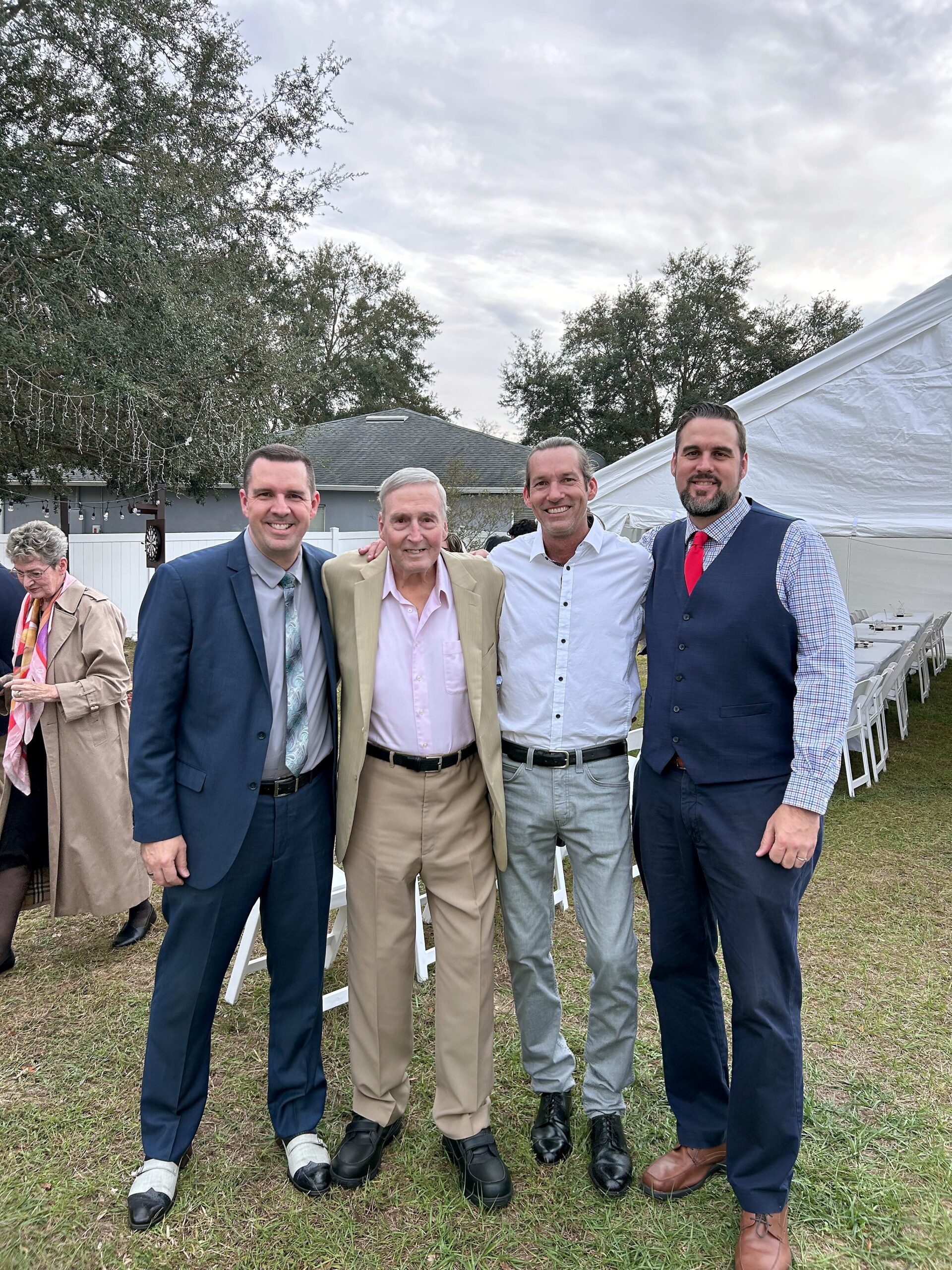 Four men in formal attire posing together at an outdoor event, with a white tent and string lights in the background.