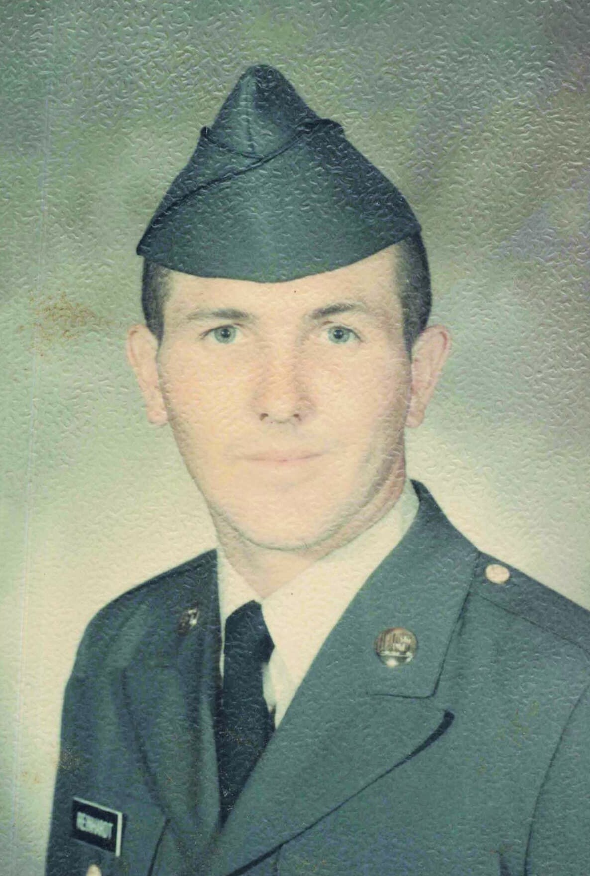 Portrait of a young man in a formal military uniform with a folded cap, wearing a tie and lapel insignia, facing the camera.