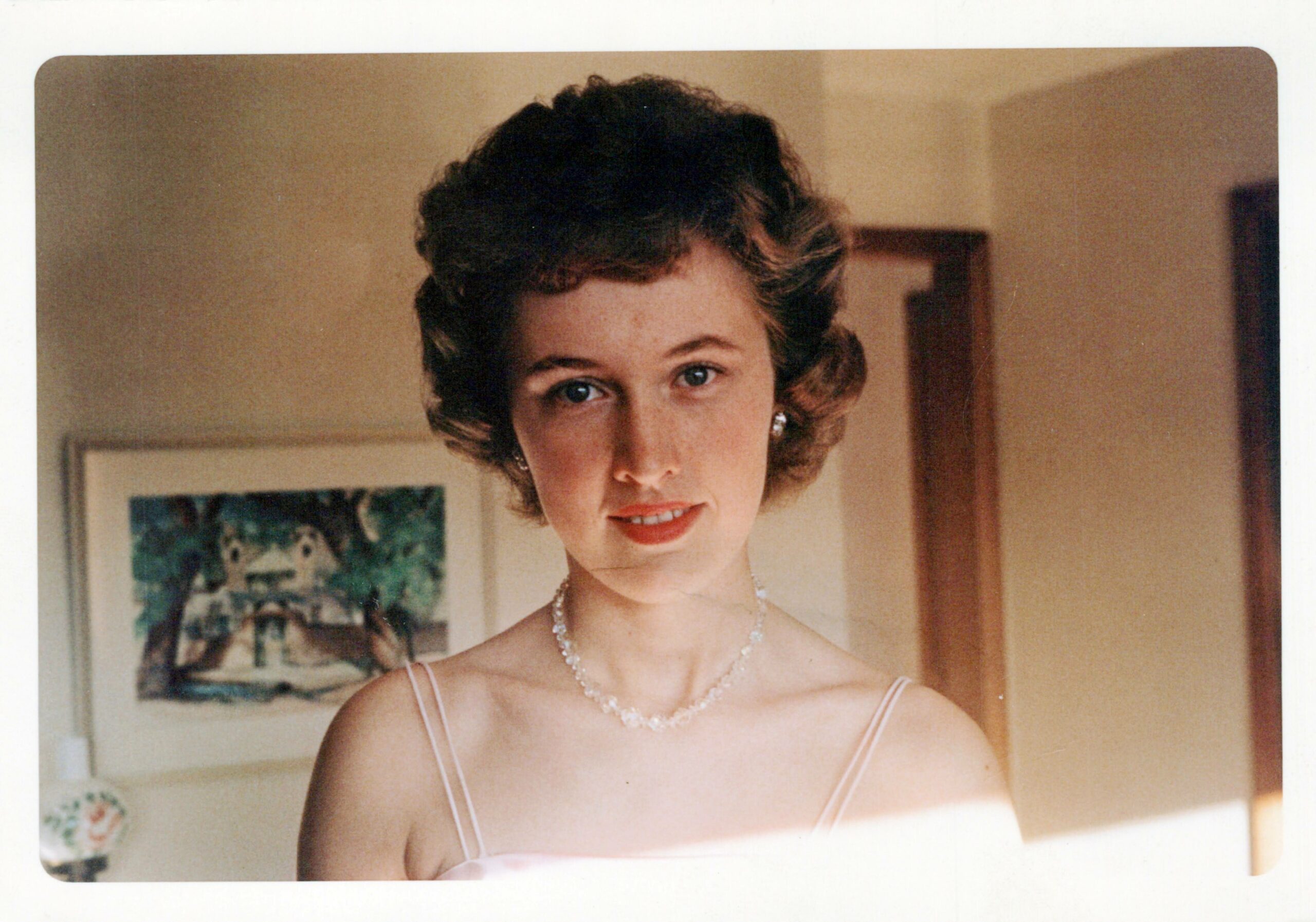 Portrait of a woman in a pale pink spaghetti-strap dress wearing a pearl necklace, indoors in a home setting.