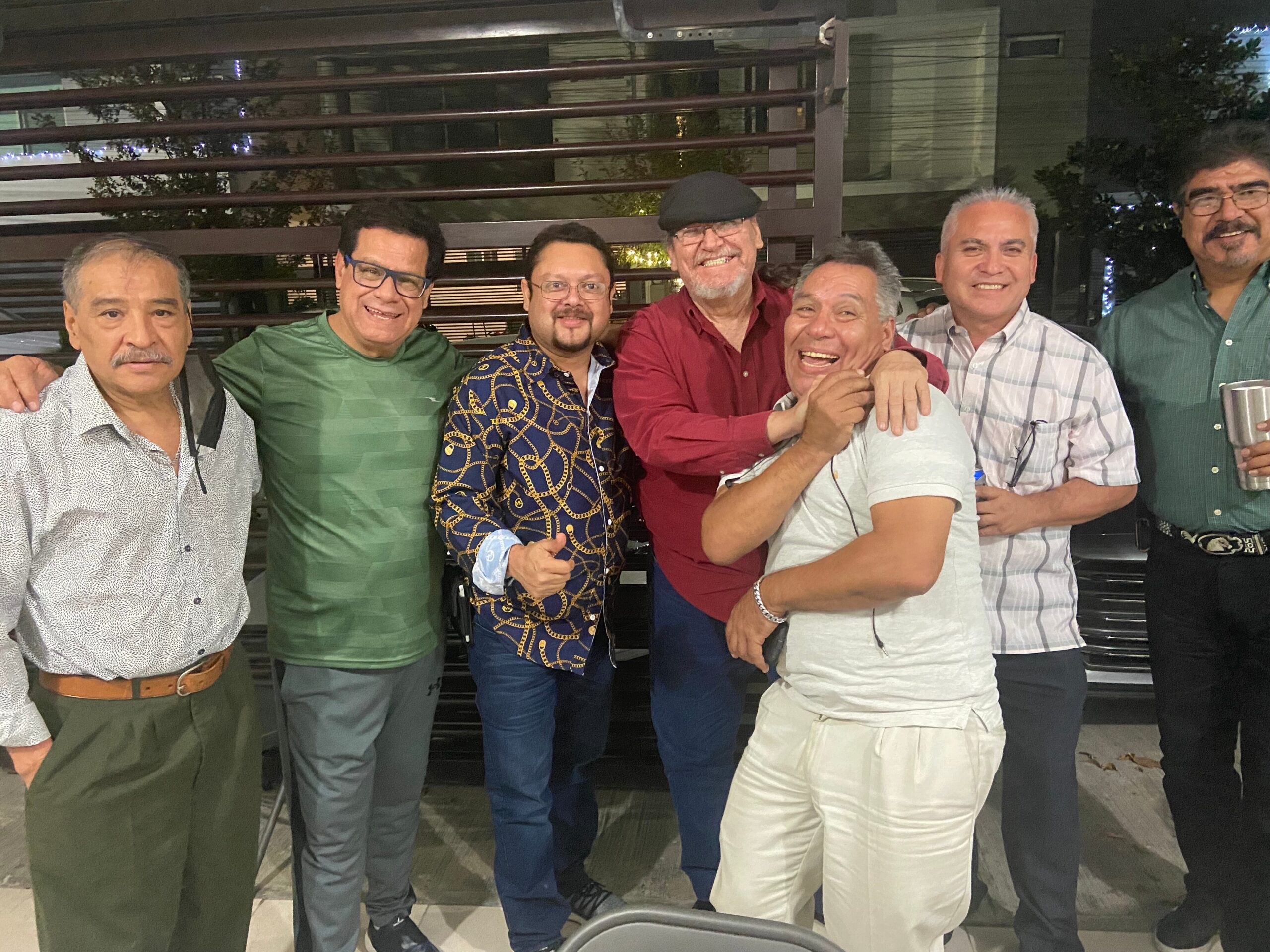 Seven men stand close together outdoors at night, smiling for the camera, with a dark railing behind them.