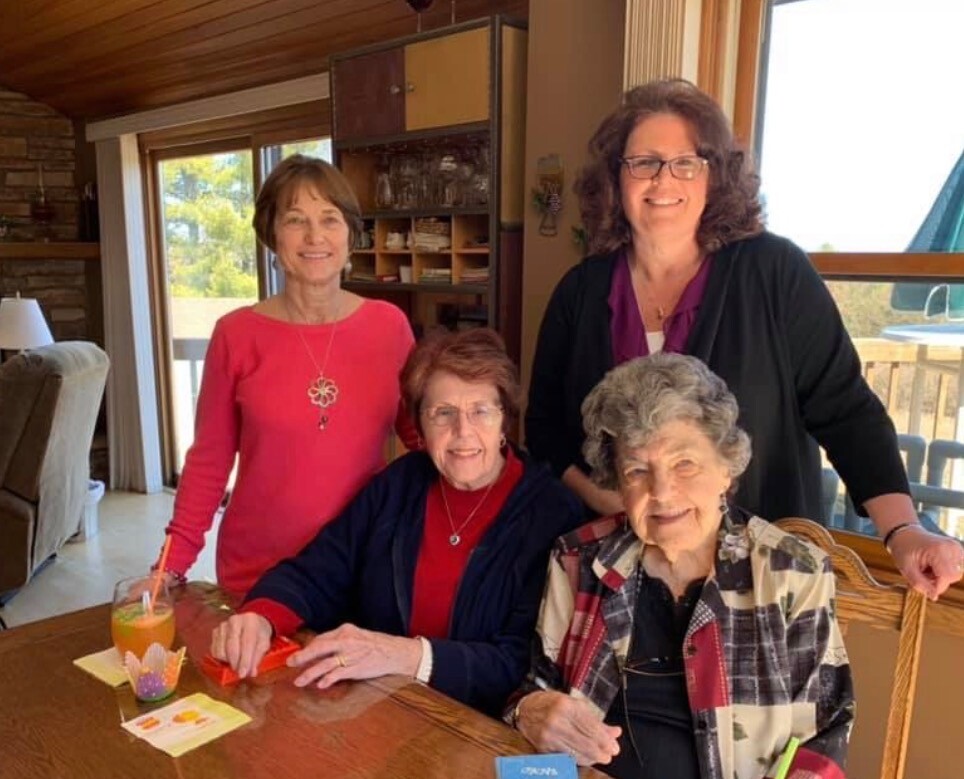 Four women of varying ages smile at the camera while gathered around a wooden dining table indoors, with a glass of orange juice and notes on the table.