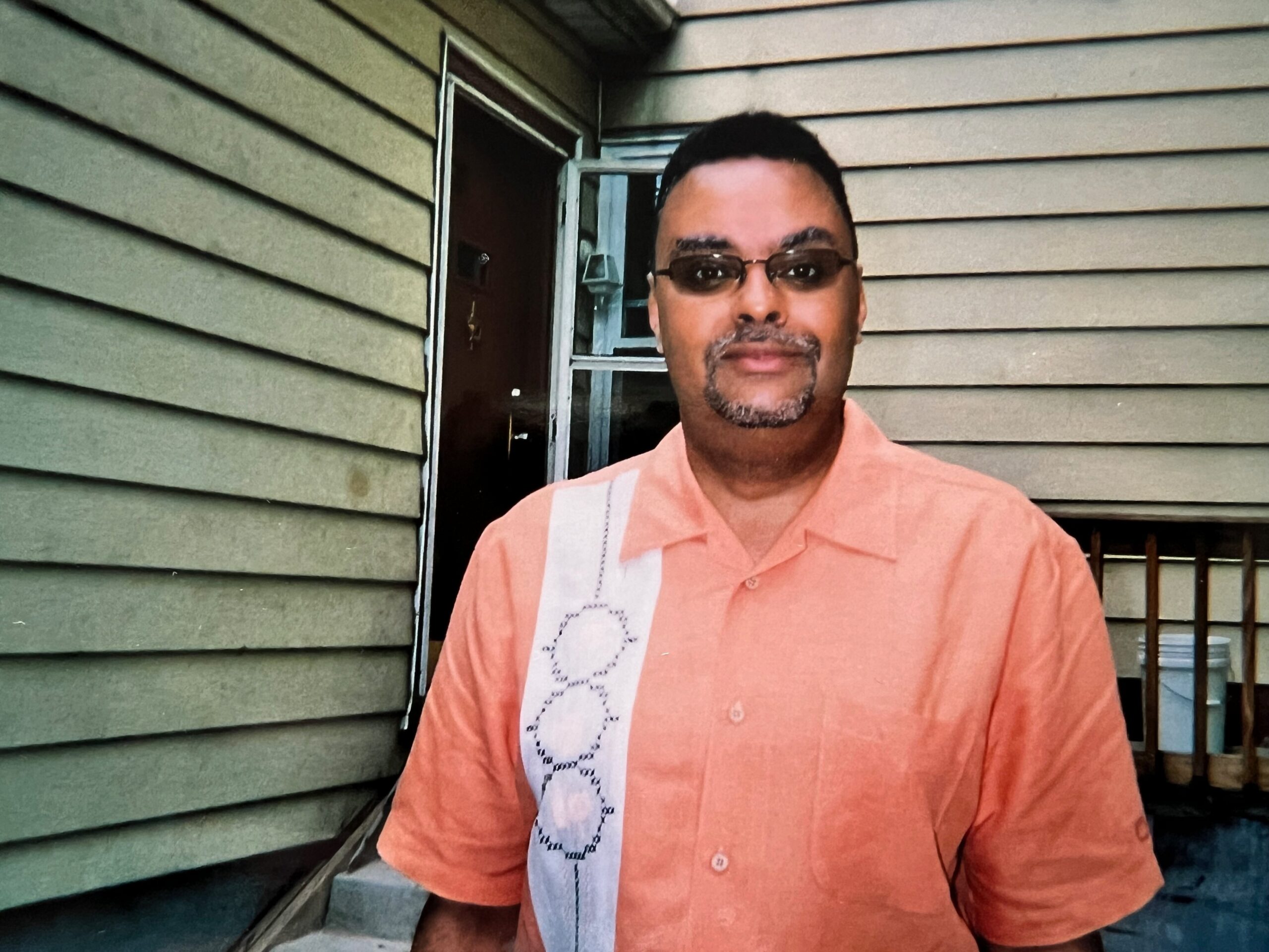 Man in an orange shirt standing outside a house with beige siding and a doorway behind him, wearing glasses and a goatee.