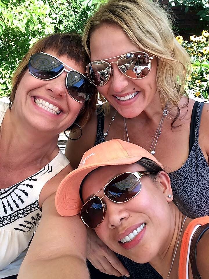 Three women wearing sunglasses pose for a sunny outdoor selfie, smiling at the camera.