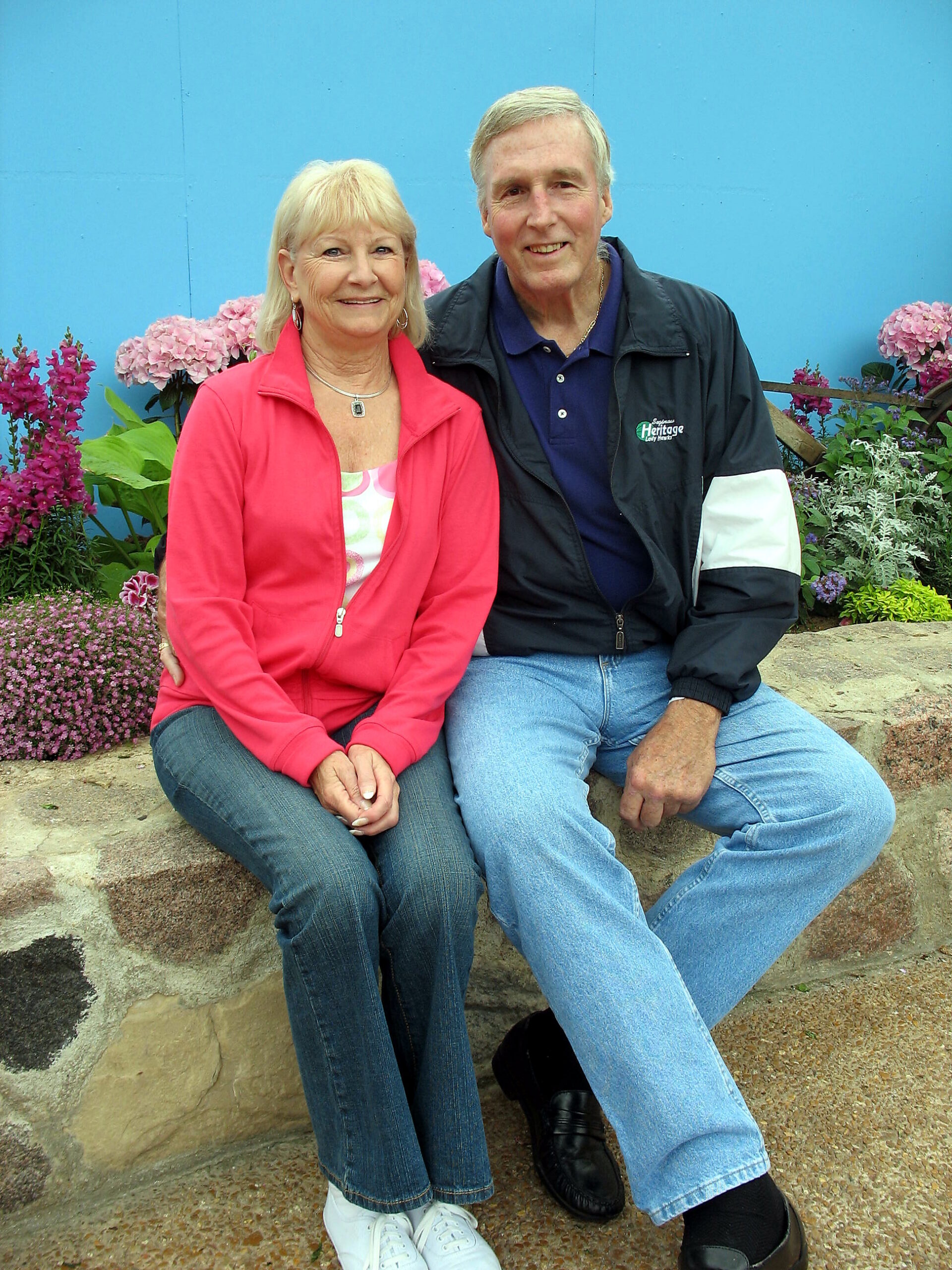An older couple sits close together on a stone wall, smiling, with a bright blue wall and flowers behind them.