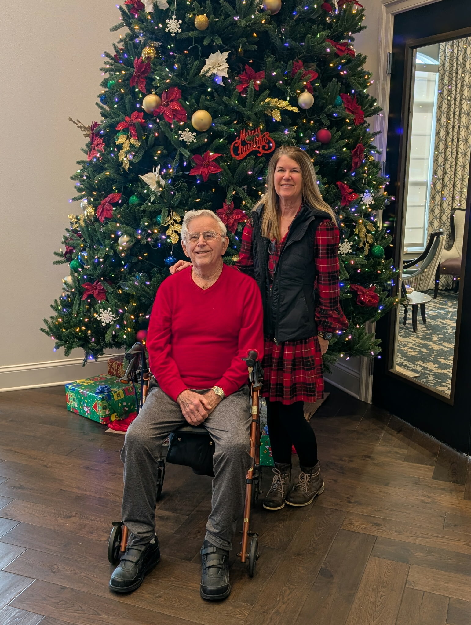 Elderly man in a red sweater in a wheelchair beside a smiling woman, posing in front of a decorated Christmas tree with gifts.