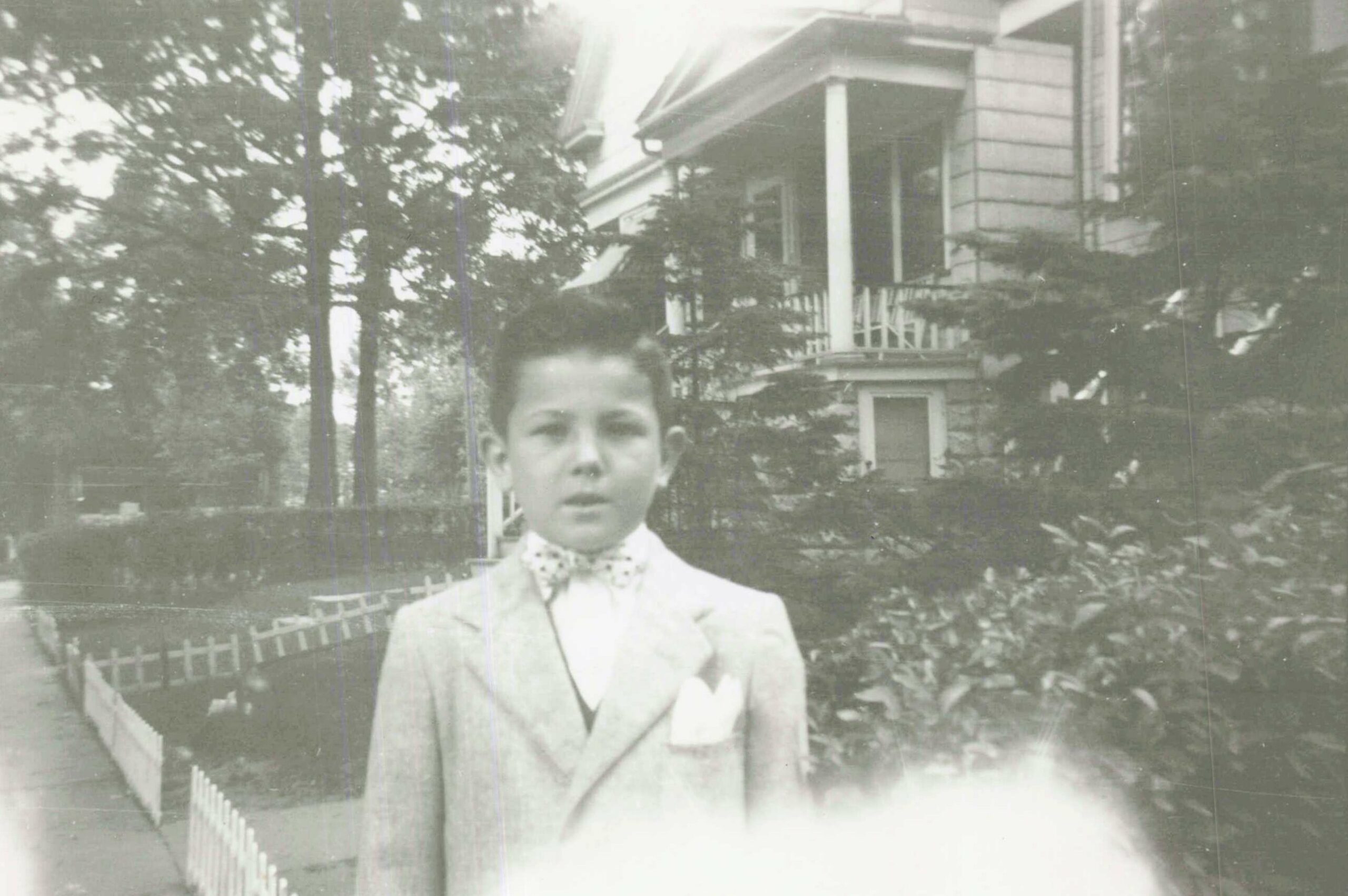 Man in a light suit standing outdoors in front of a two-story house with a porch.