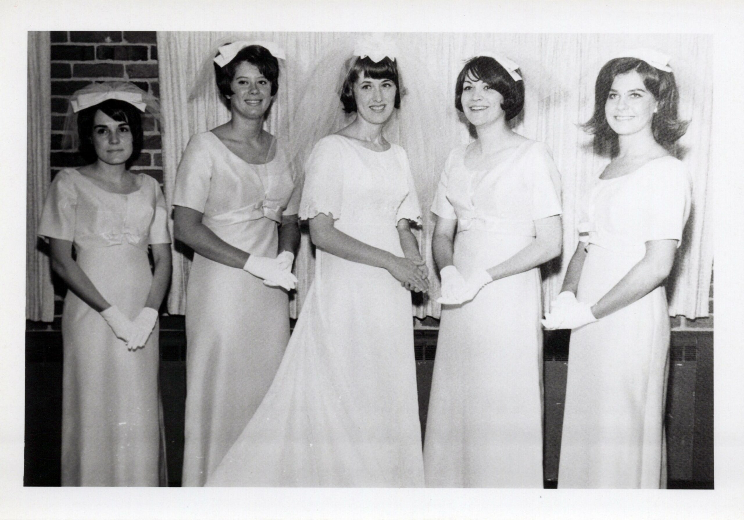 Five women in white uniforms with gloves and bows stand in a line, shaking hands in the center of the group.