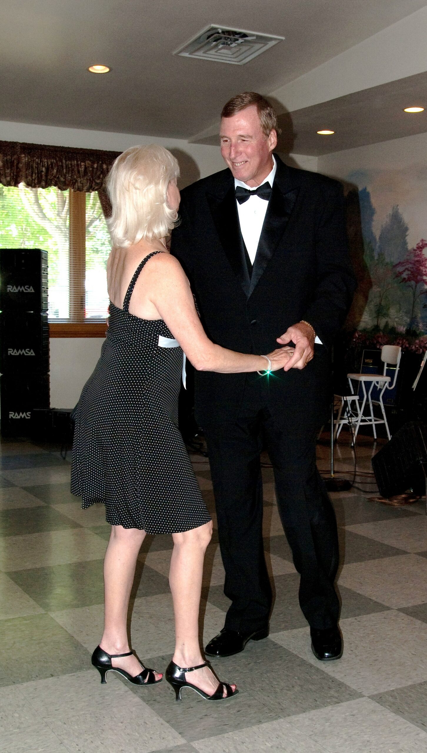 Smiling couple dancing at a formal event: woman in a black polka-dot dress and heels, man in a tuxedo.