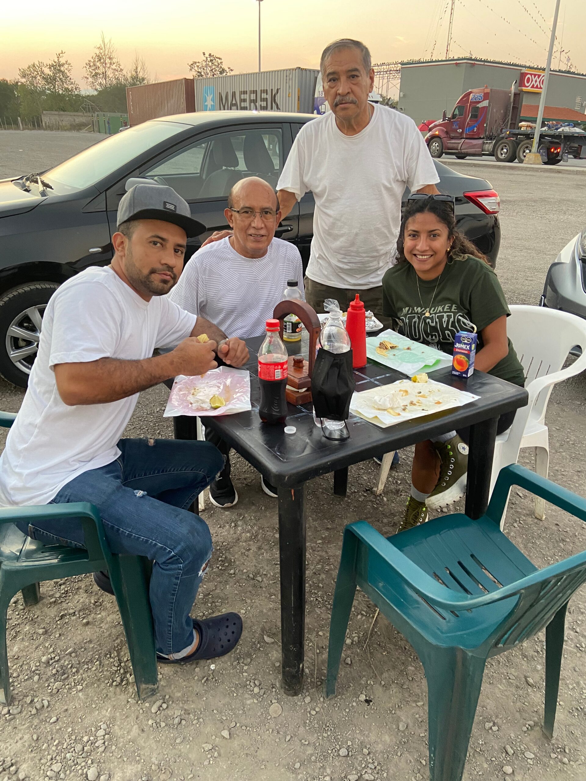 Four people share a casual meal at a plastic table in a gravel parking lot, with a parked car and trucks in the background at sunset.