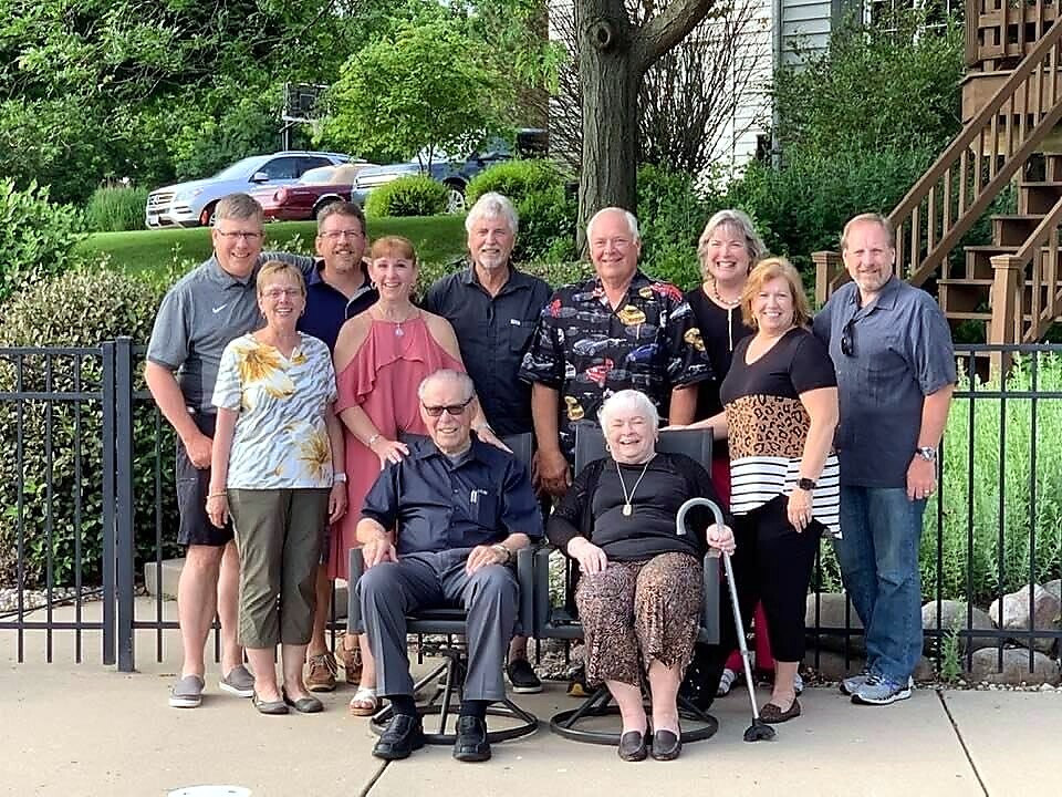 A diverse group of adults posing outdoors in front of a fence, with two elderly individuals seated in chairs at the front.