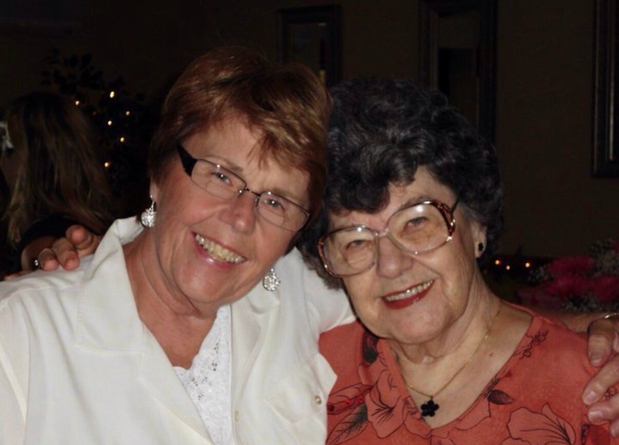 Two smiling older women with short hair and glasses posing together at a social event, one in white and the other in orange.