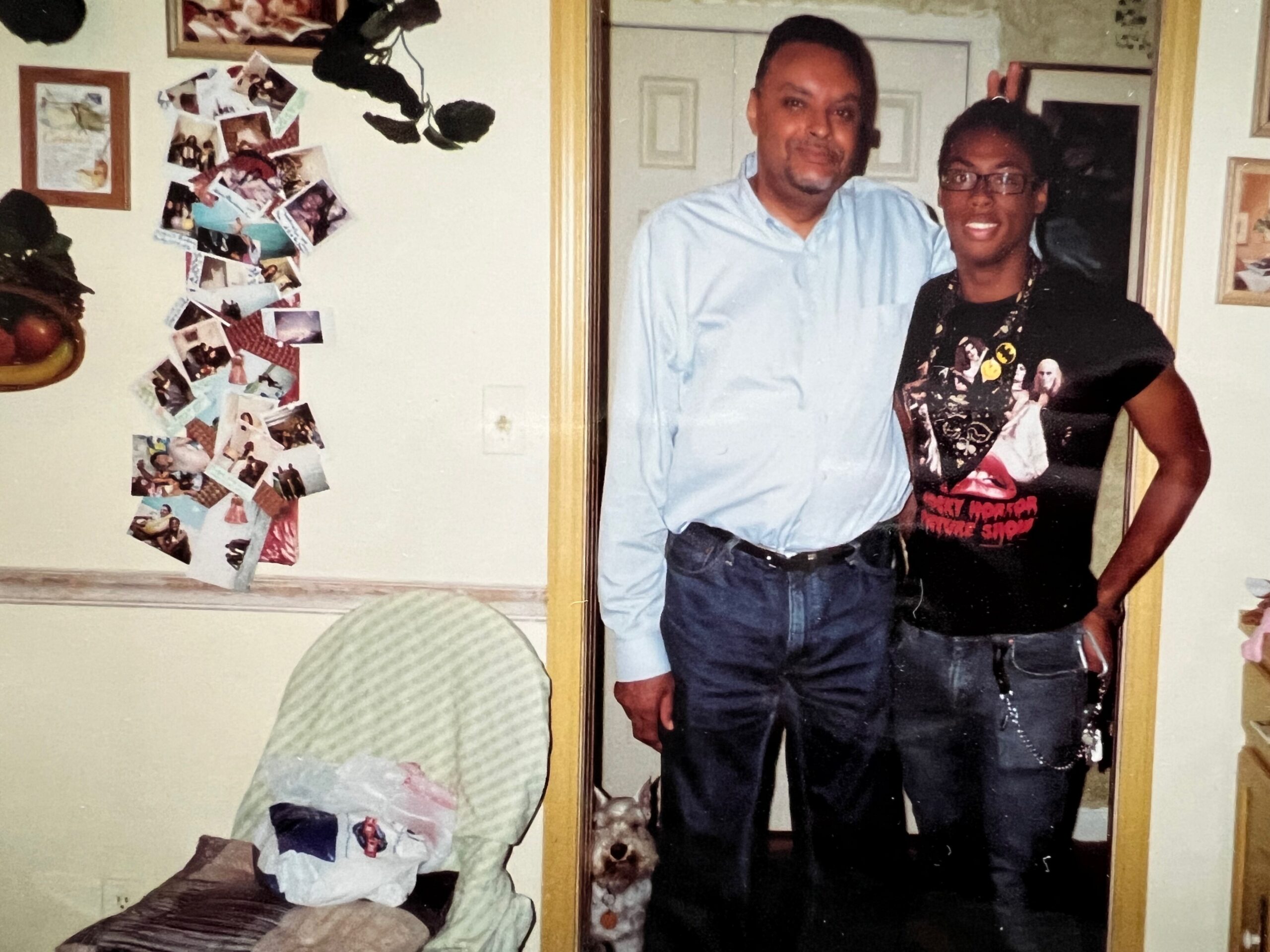 Two people pose together in a doorway, smiling, with a collage of photos on the wall to the left.