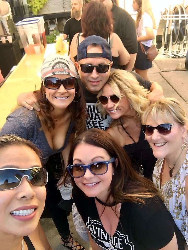 Group of six adults smiling and posing for a selfie at an outdoor event, all wearing sunglasses and casual outfits.