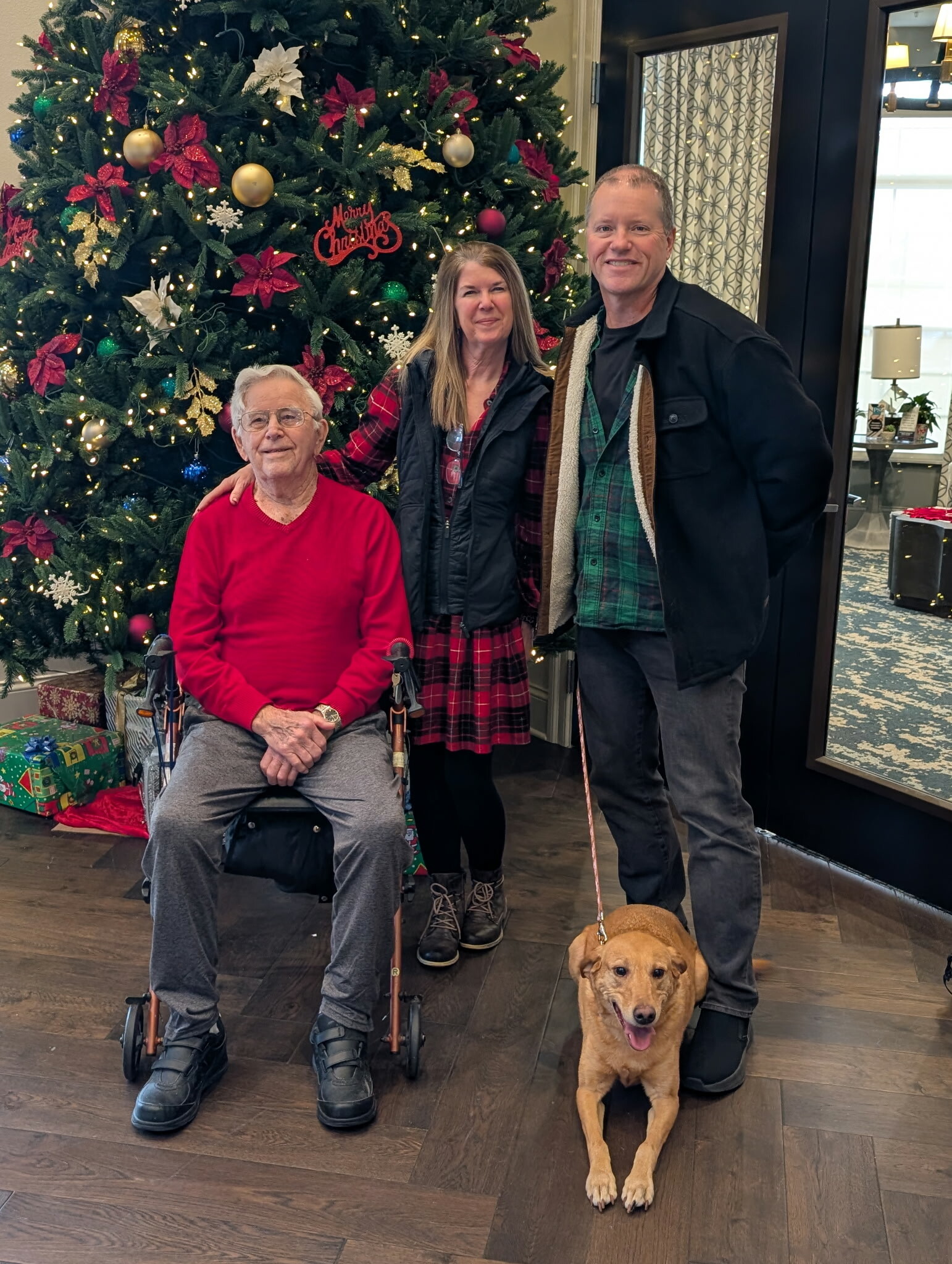 Three adults and a golden retriever posing by a Christmas tree with lights and wrapped presents nearby in a festive lobby setting.