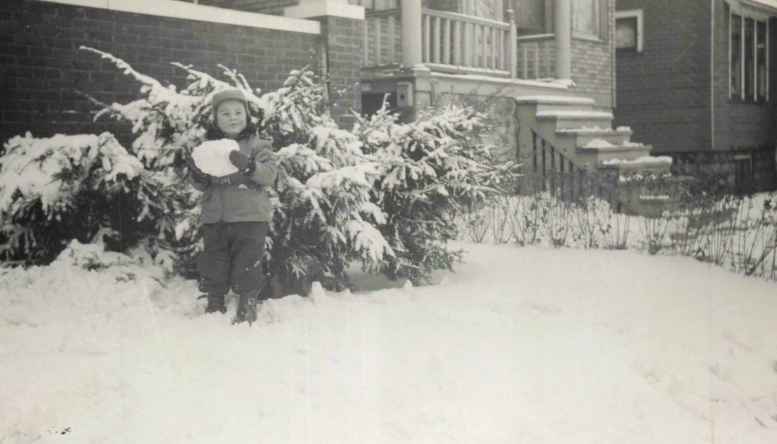 Child in a winter coat and hat holding a snowball in a snow-covered front yard, with a brick house and snow-laden shrubs in the background of a residential street.