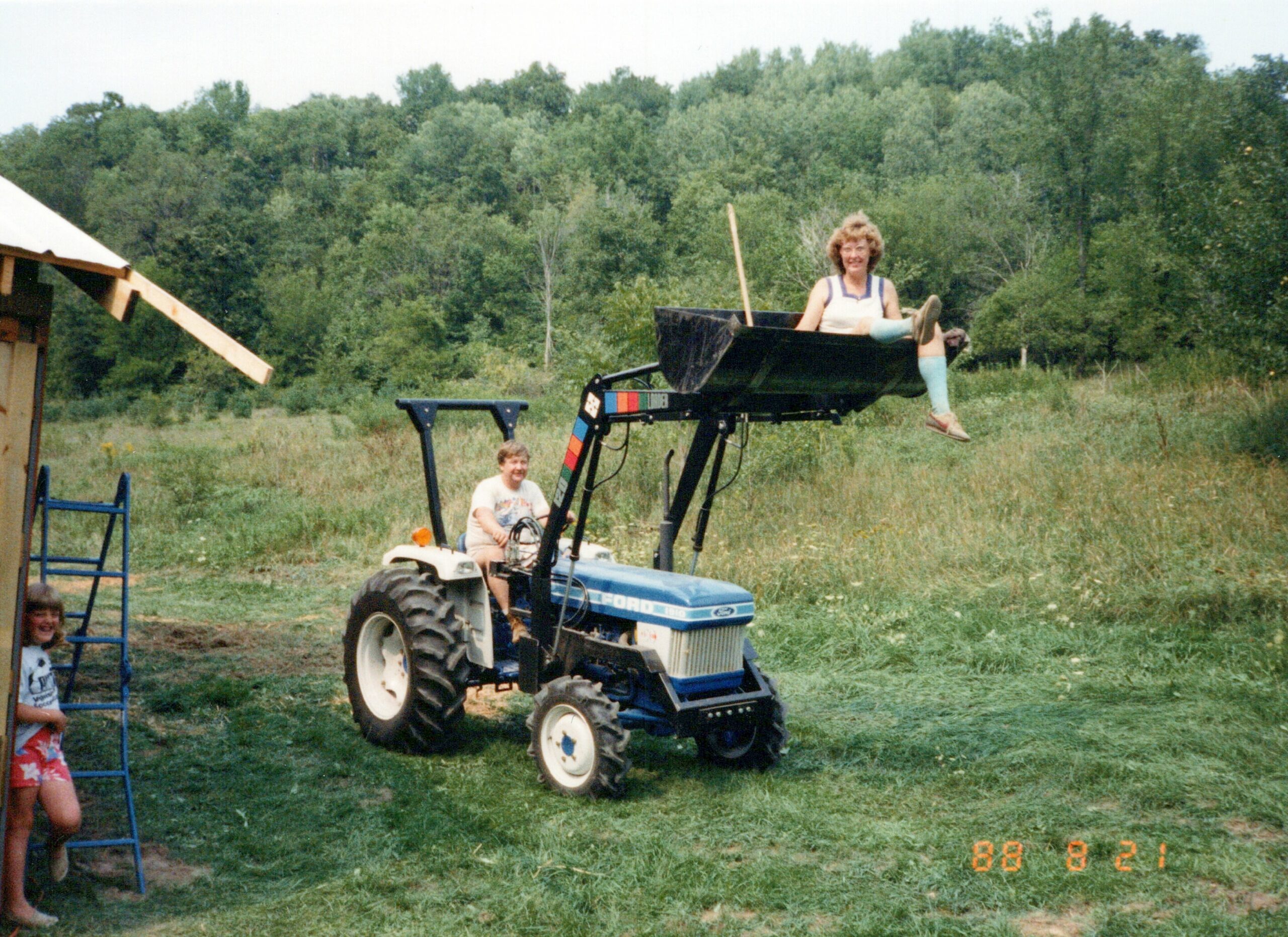 Farm scene: a blue tractor with a raised front loader, person sitting in the bucket with legs dangling off the edge, others nearby in a grassy field.