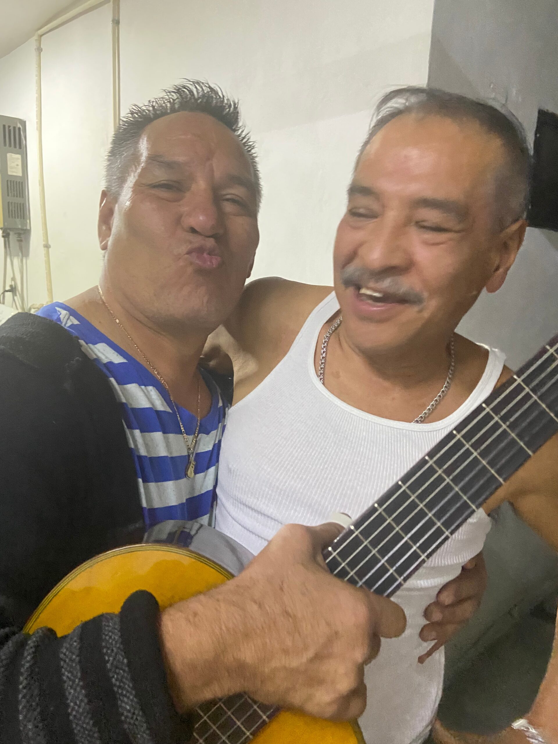 Two men smiling for a selfie, one in a striped blue shirt and the other in a white tank top holding a guitar.