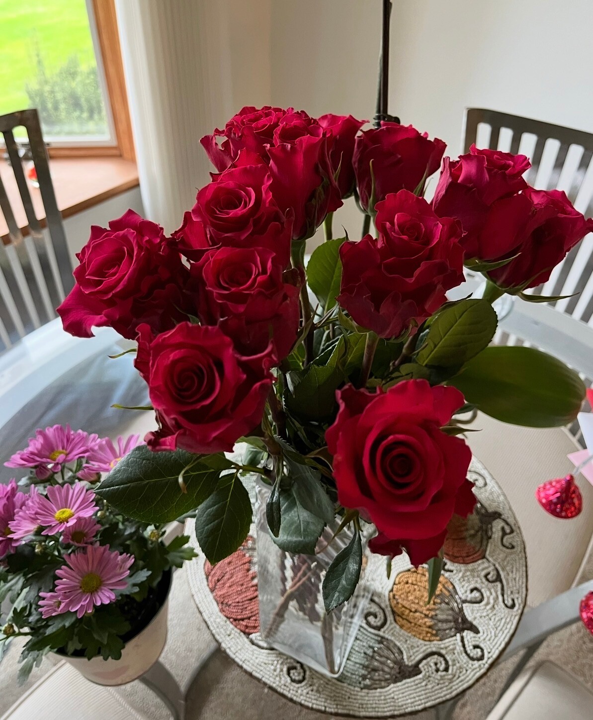 Bouquet of deep red roses in a glass vase on a patterned centerpiece table indoors, with pink daisies in a pot nearby.