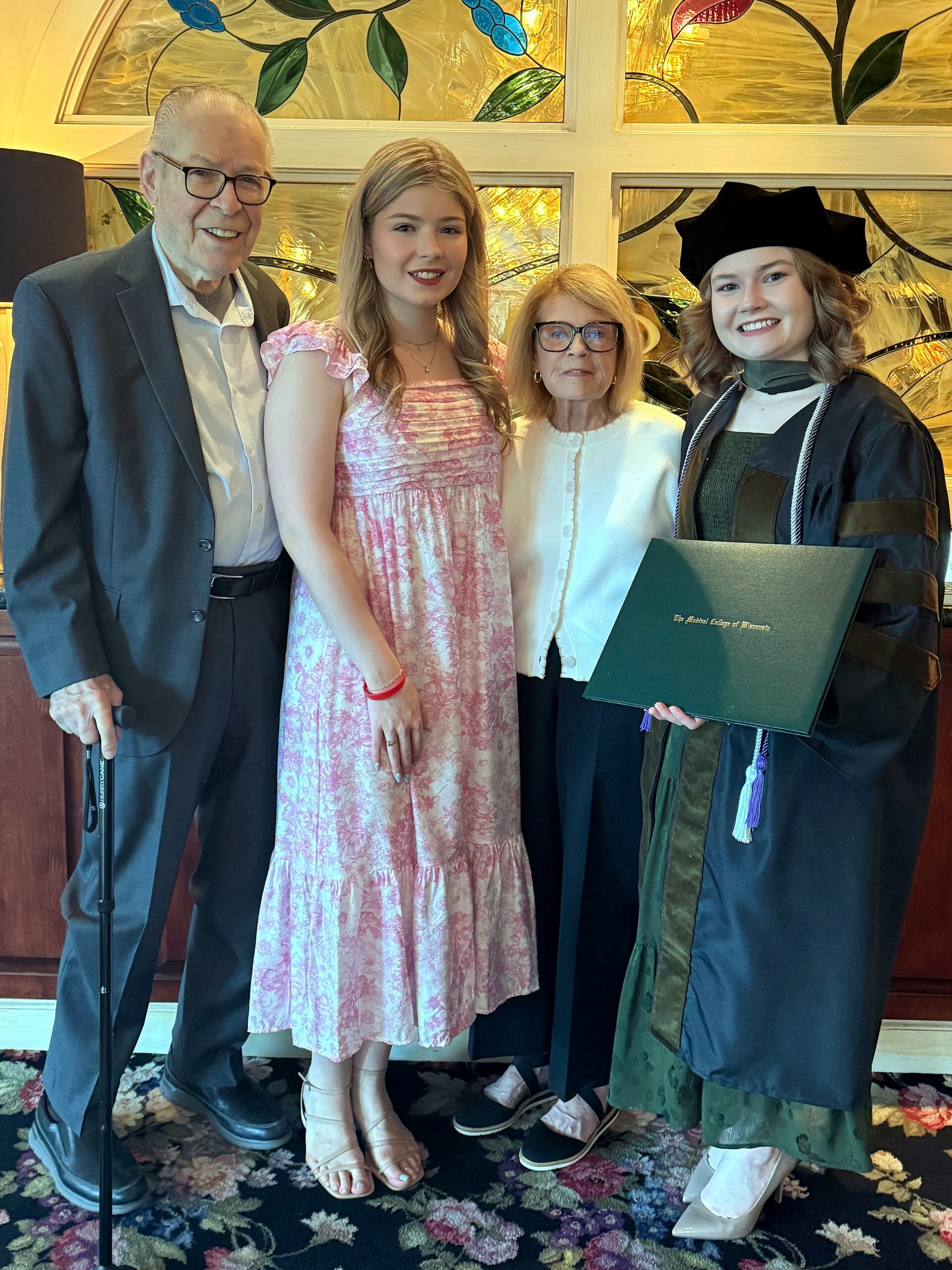 Four people posing for a graduation photo: an elderly man in a suit, a young woman in a pink patterned dress, an older woman with glasses in a white cardigan, and a smiling graduate in a cap and gown holding a green diploma cover.