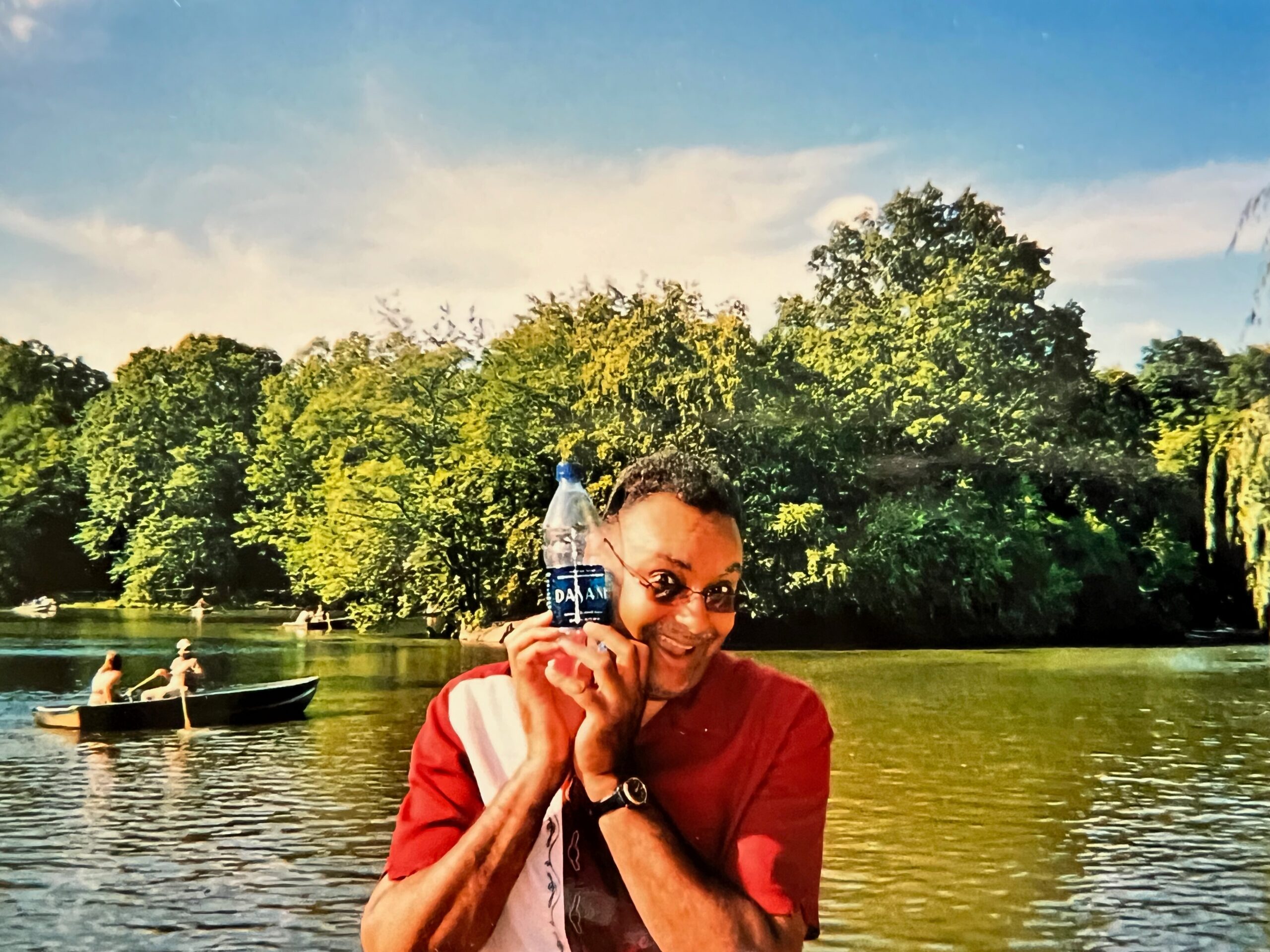 Smiling man in a red shirt holds a water bottle up to his face by a lake, with trees and boats in the background.