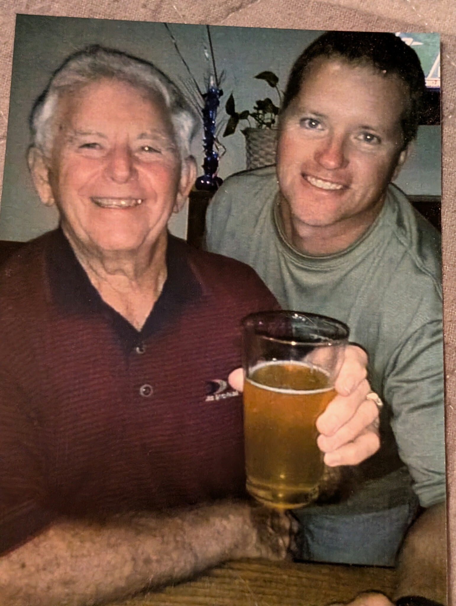 Two smiling men posing together, an older man on the left and a younger man on the right, the younger holding a pint of beer.