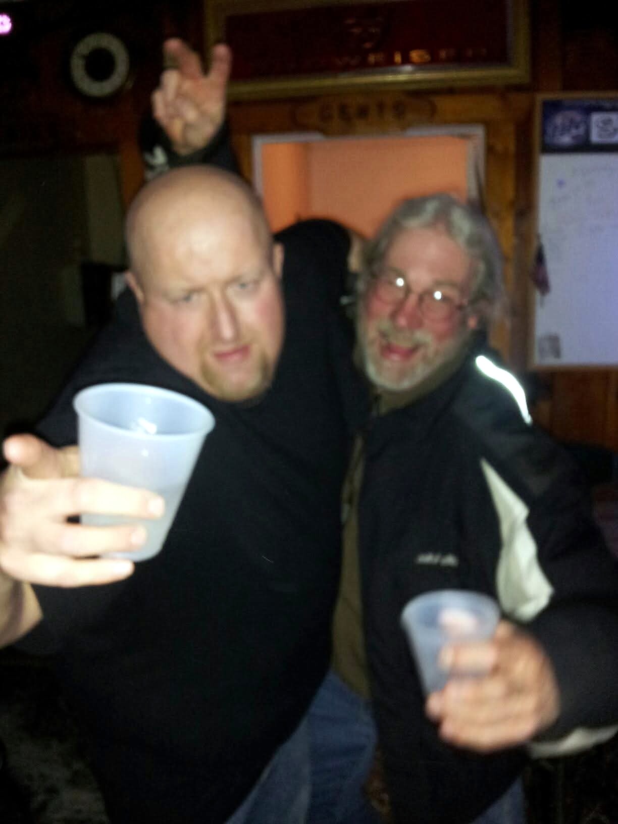 Two men raise plastic cups in a friendly toast at a dimly lit bar; foreground is a bald man in black shirt.