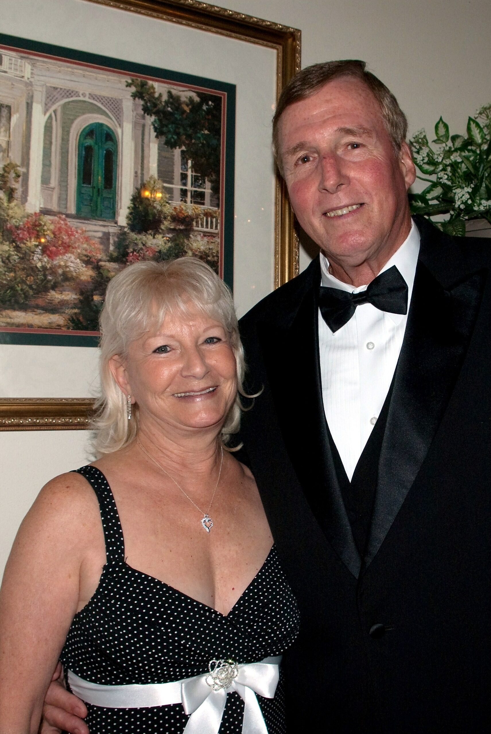 Smiling couple in formal attire pose together indoors, woman in polka-dot dress with white ribbon, man in tuxedo.