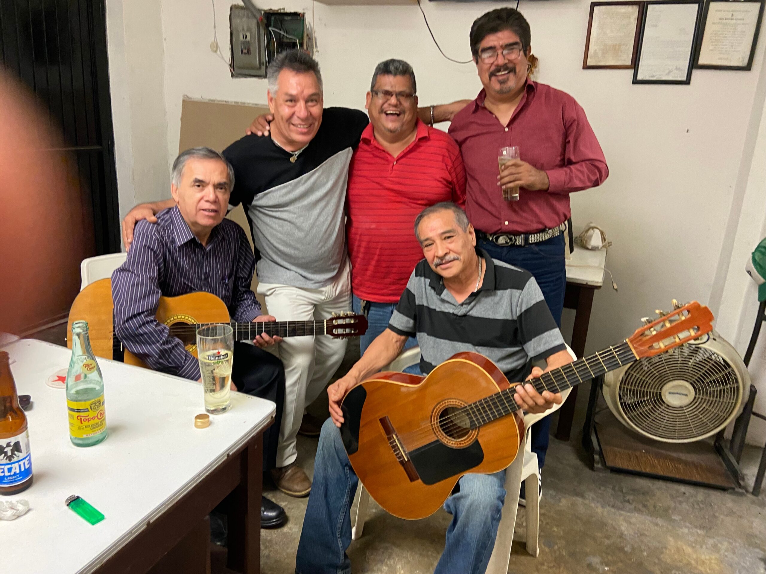 Five men posing together in a room; two hold acoustic guitars and drinks are on the table.