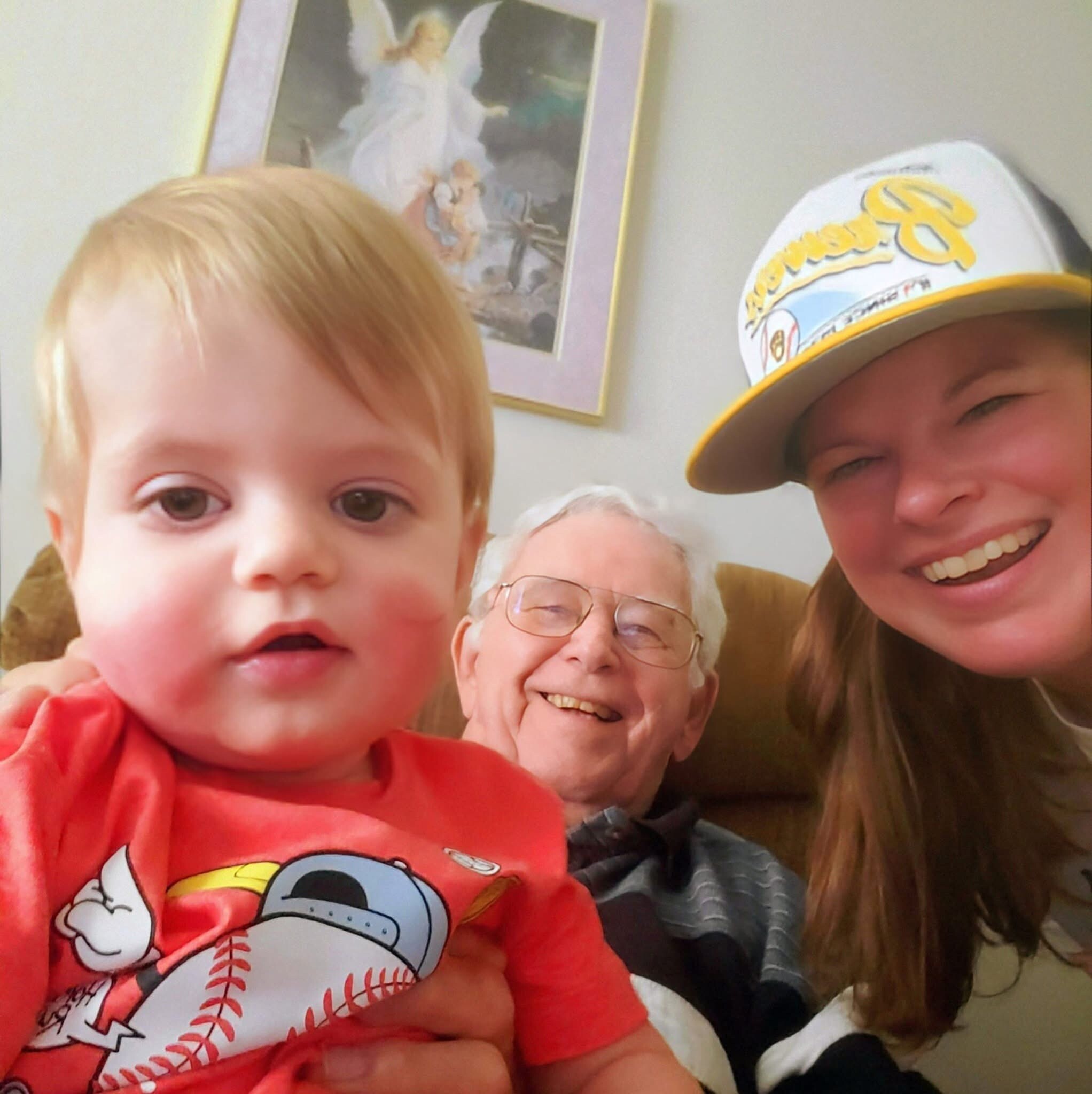 Close-up group selfie: a young child in a red shirt with a baseball graphic, an older person with glasses, and another person wearing a yellow cap indoors.