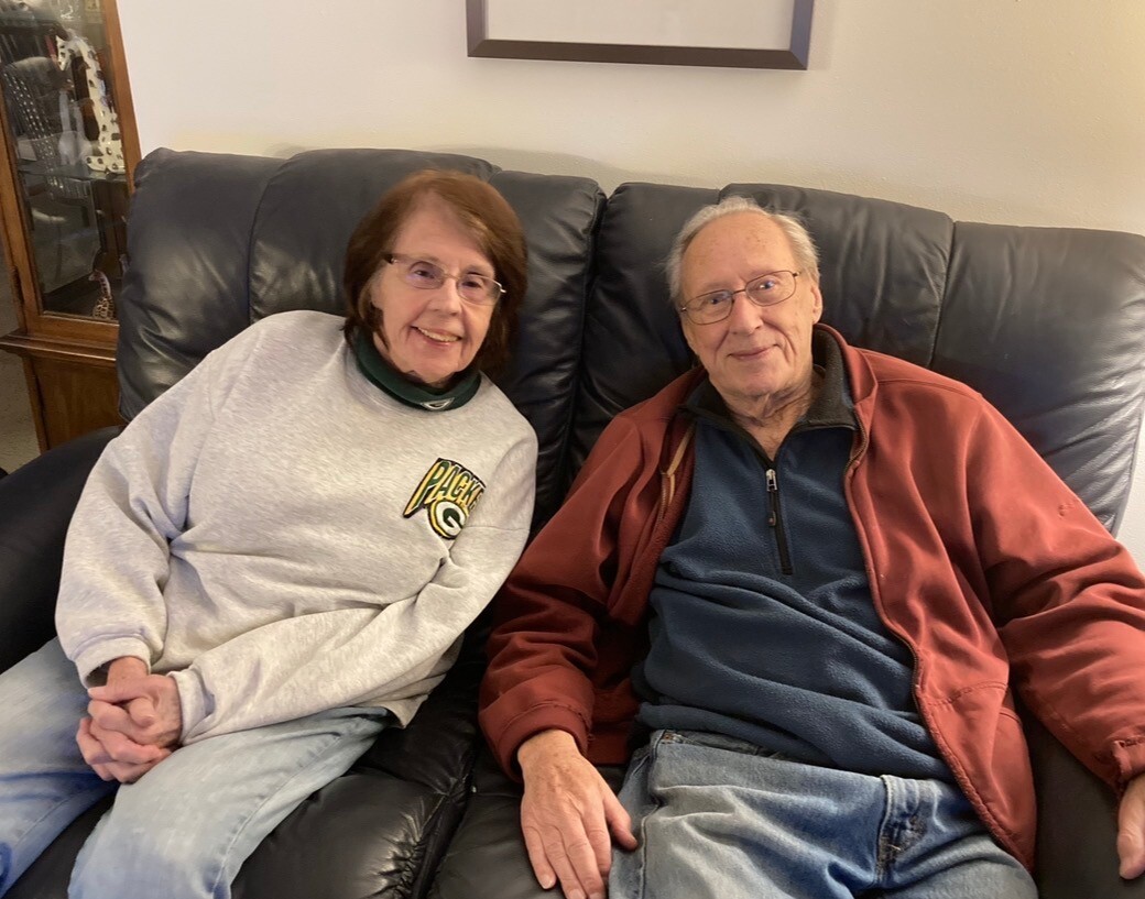 Two elderly adults sit close together on a black leather couch, smiling at the camera in a cozy living room. The person on the left wears a light gray Packers sweatshirt and light jeans, while the person on the right wears a red jacket over a navy fleece and jeans.