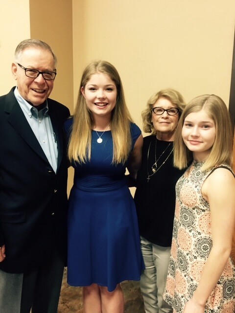 Group photo of four people posing together indoors: an older man in a blazer, two young women in blue dress and patterned sleeveless dress, and an older woman in black top with glasses.