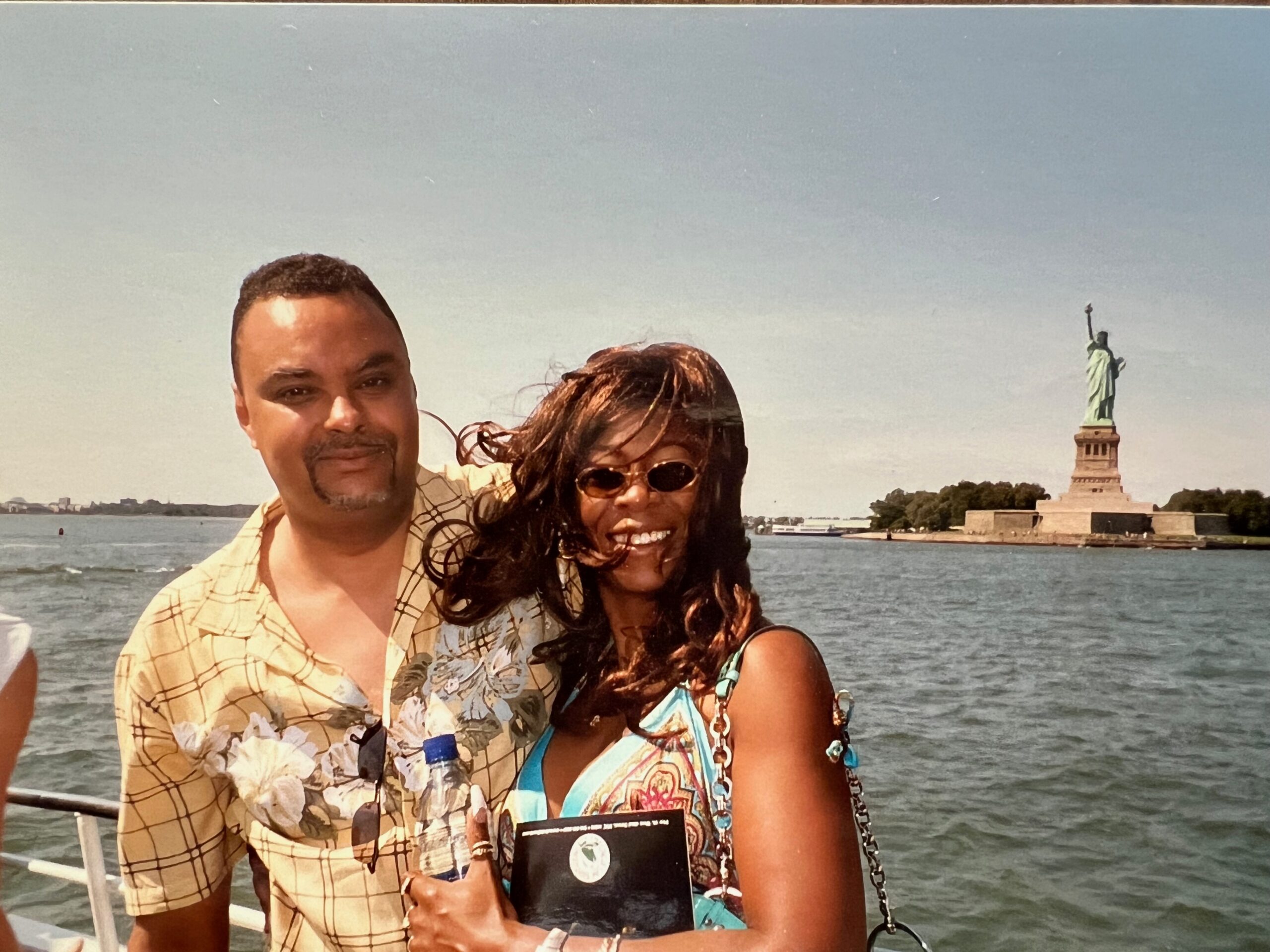 Couple posing on a boat with the Statue of Liberty in distance over the water on a sunny day
