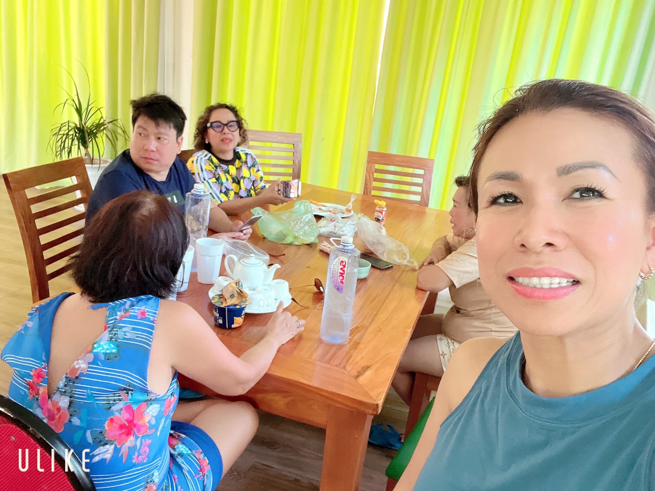 Smiling woman takes a selfie at a wooden dining table with friends seated around, bright lime curtains in the background.
