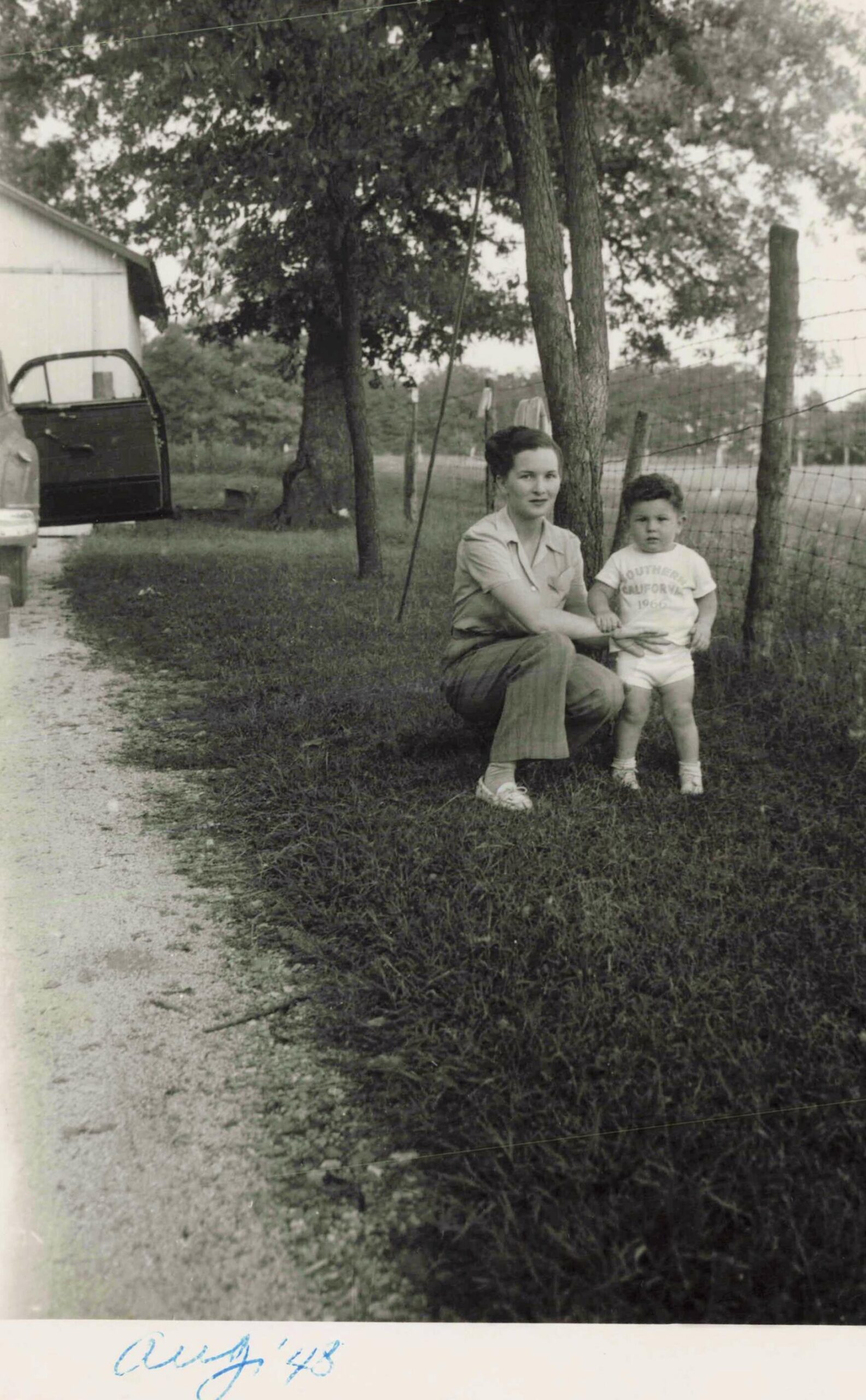 Woman kneels beside a young child outdoors in a yard with trees and a wire fence, near a parked car with its door open (black-and-white photo).
