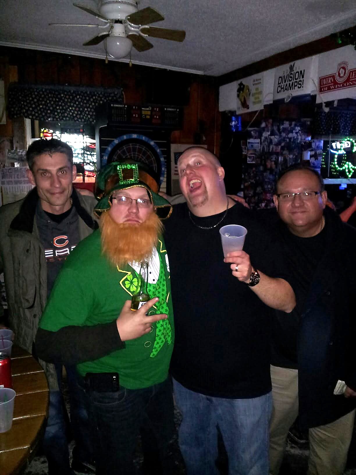 Group of four men inside a dim bar, one wearing a green leprechaun outfit with a beard posing with drinks and friends nearby.