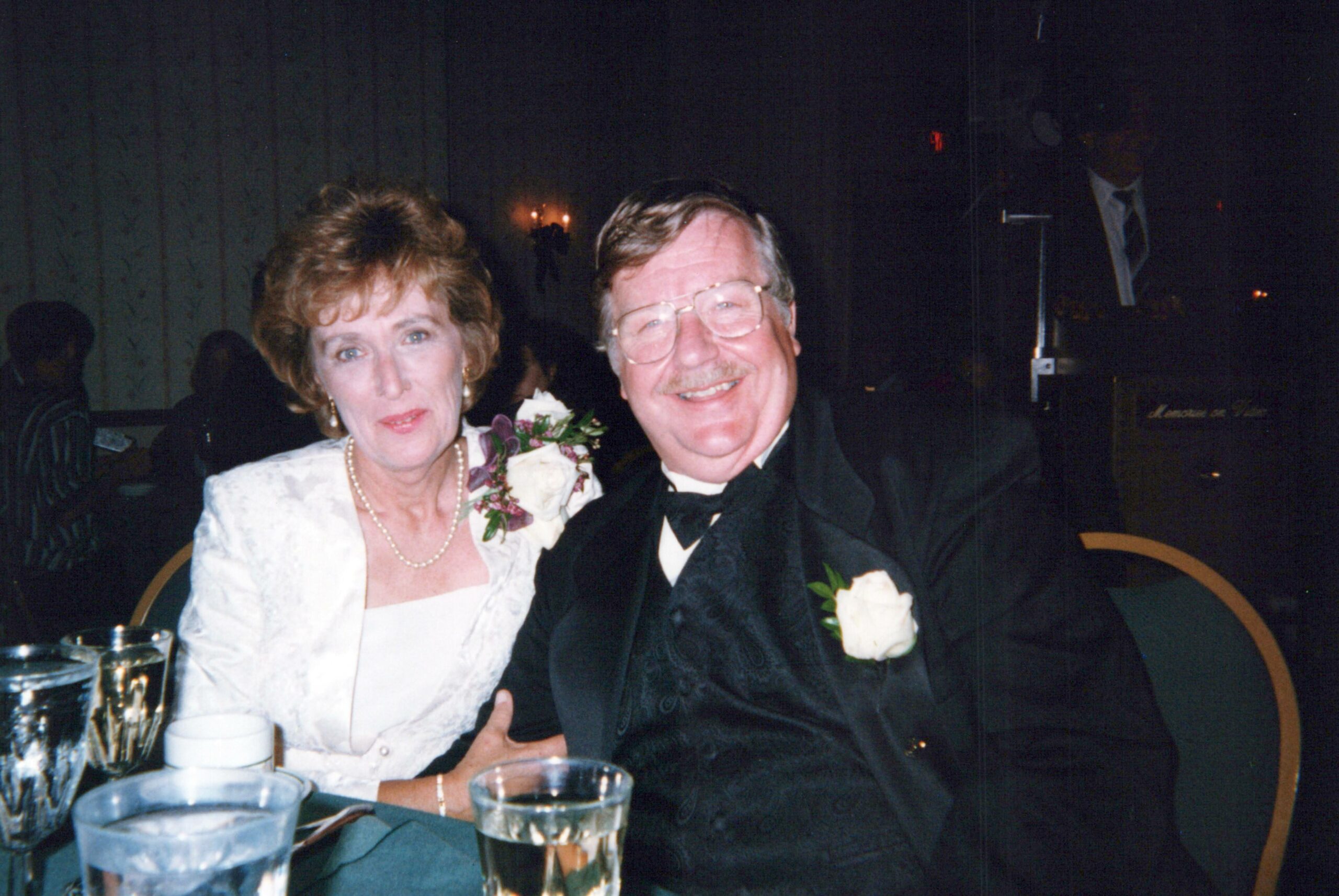 Smiling older couple in formal attire at a dimly lit event, seated at a table with drinks, likely a wedding reception.