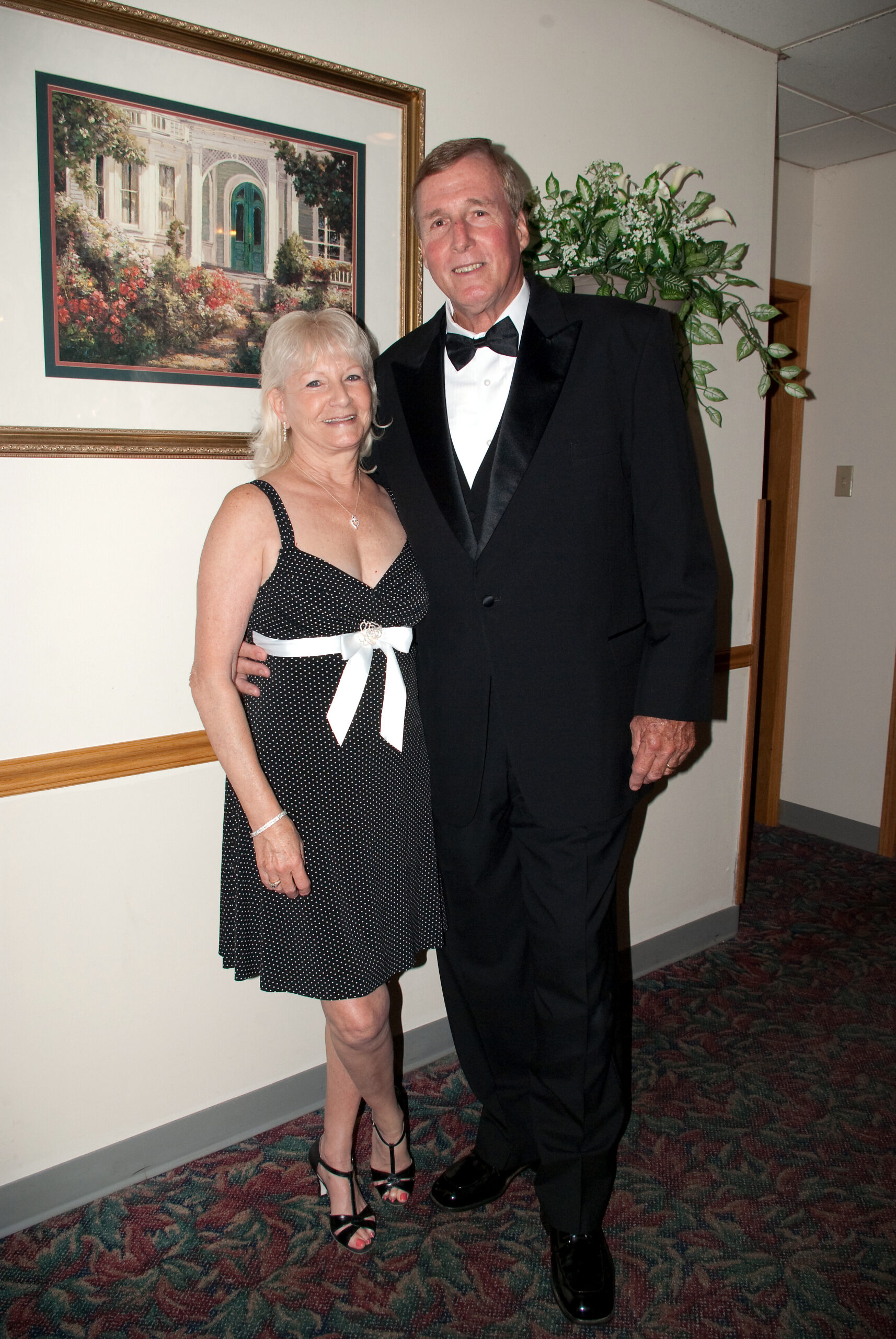 Smiling couple in formal attire posing indoors; woman in a black polka-dot dress with a white sash and heels, man in a black tuxedo, framed painting behind them.