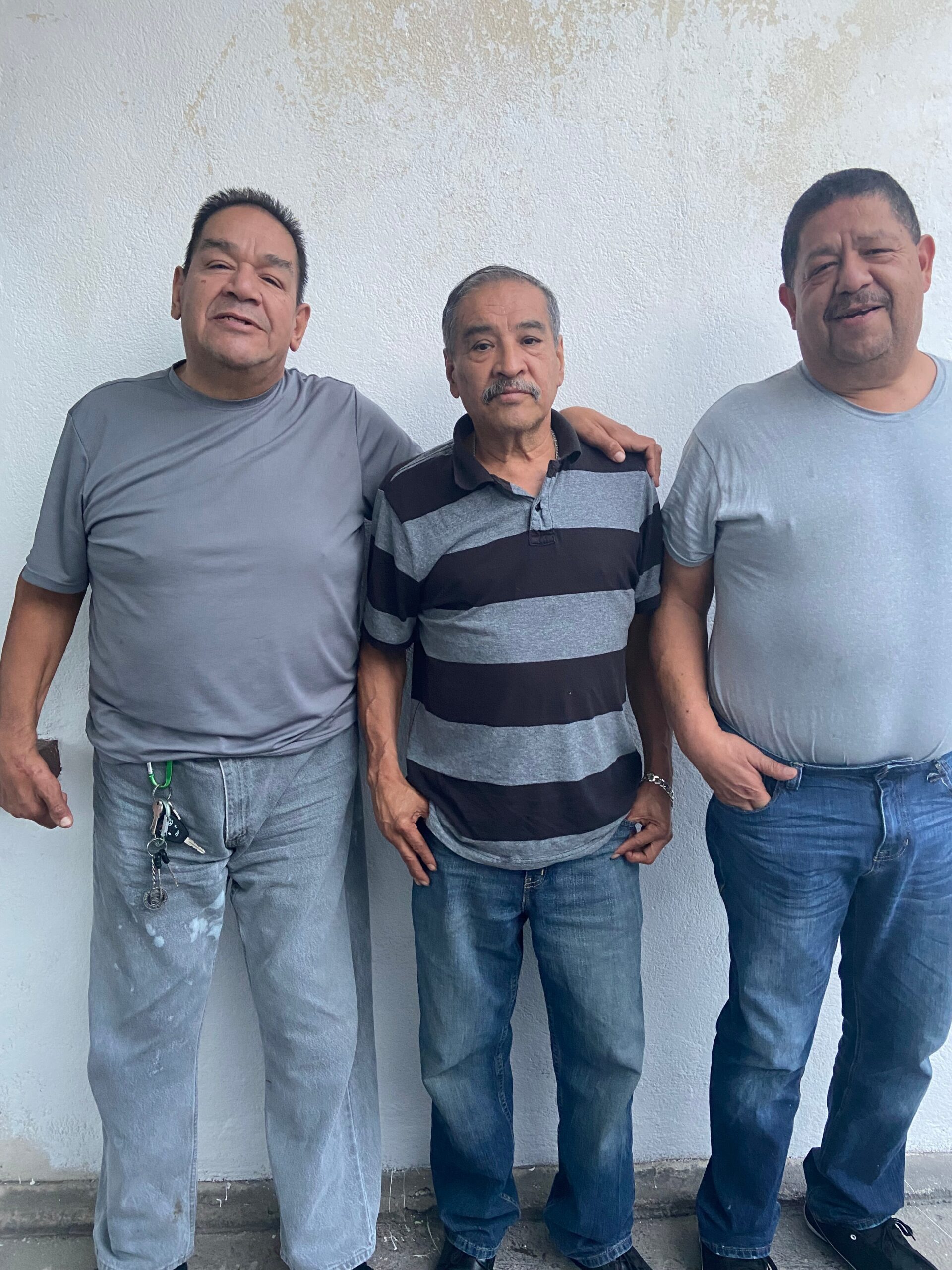 Three men standing side by side against a white, worn wall with their arms around each other's shoulders, casual clothing.