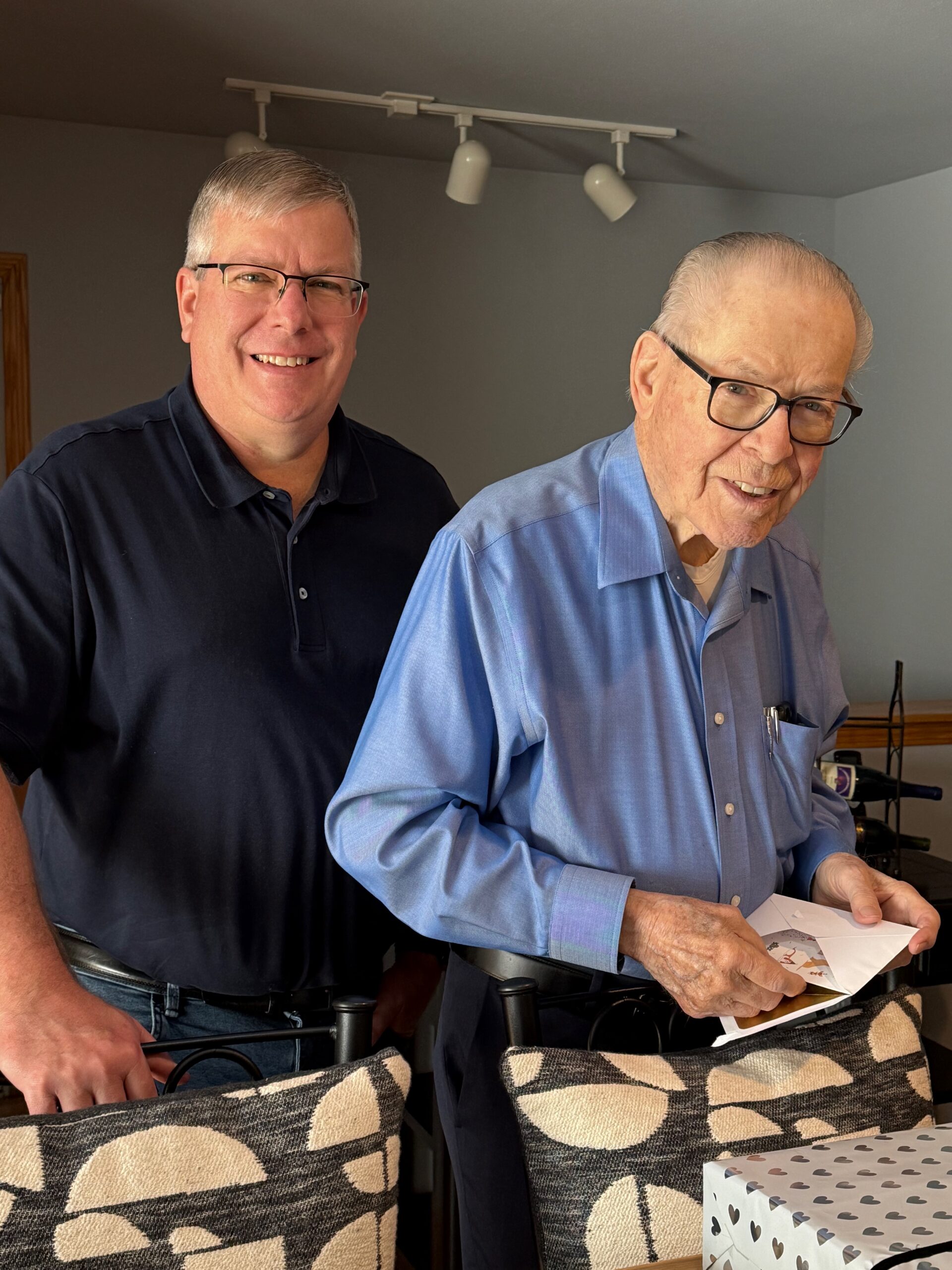 Two men indoors, one younger in a dark polo smiling beside an older man in glasses who is cutting paper at a table with gift boxes nearby.