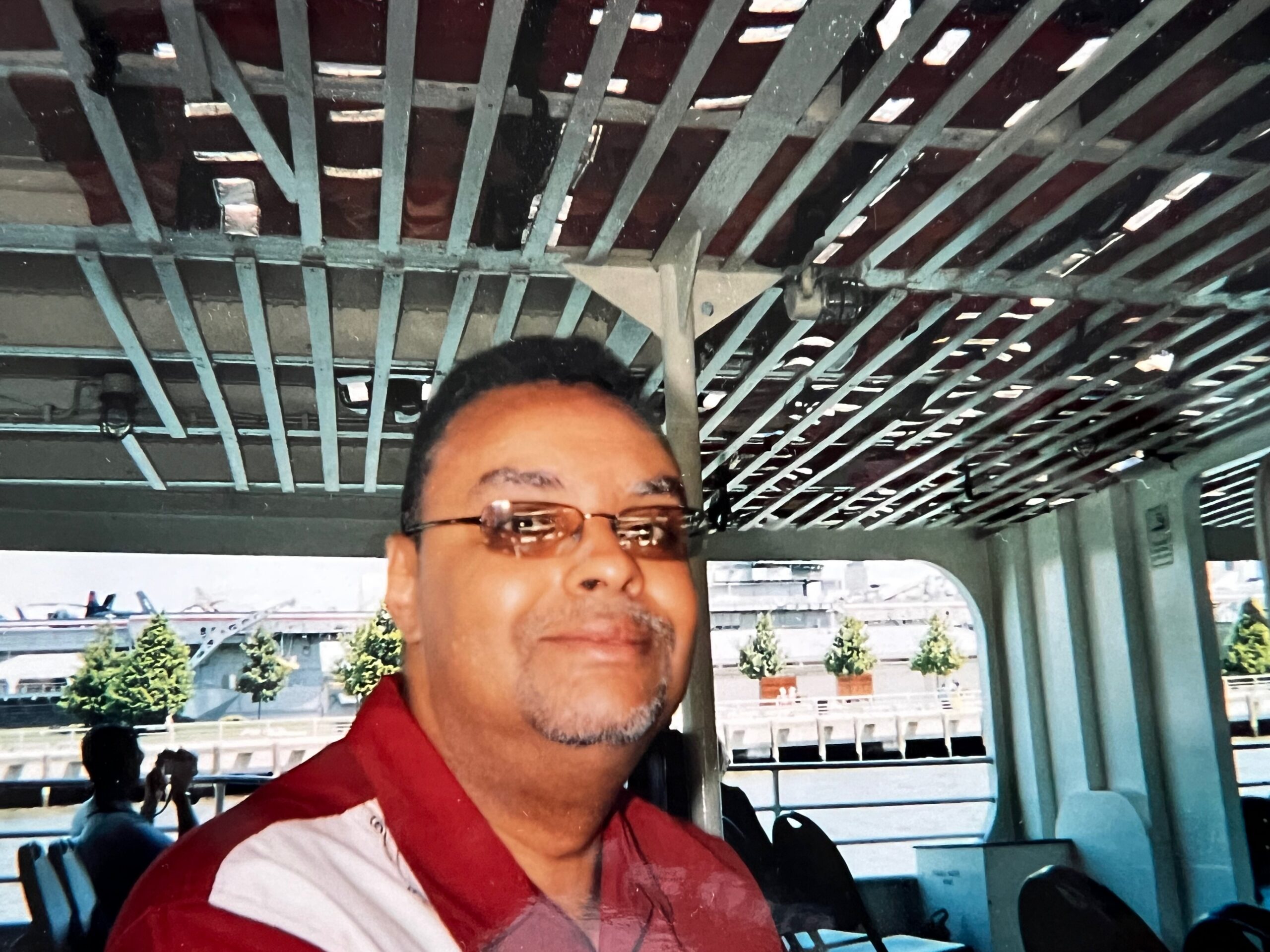 Close-up of a man wearing glasses and a red-and-white shirt, with a metal overhead-beam ceiling in the background.