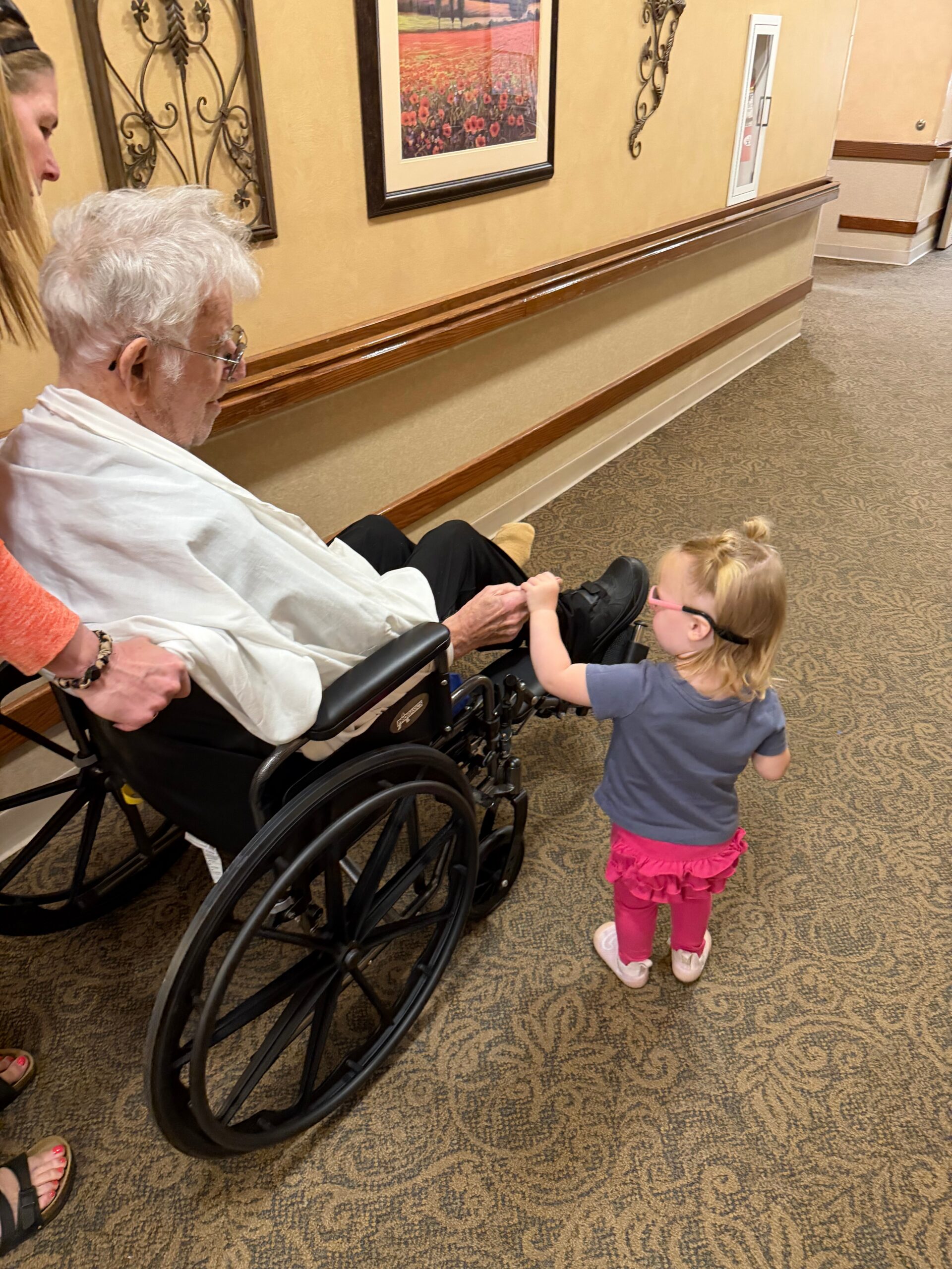 A young girl helps an elderly man in a wheelchair in a beige-hued hallway of what looks like a care facility or hotel.