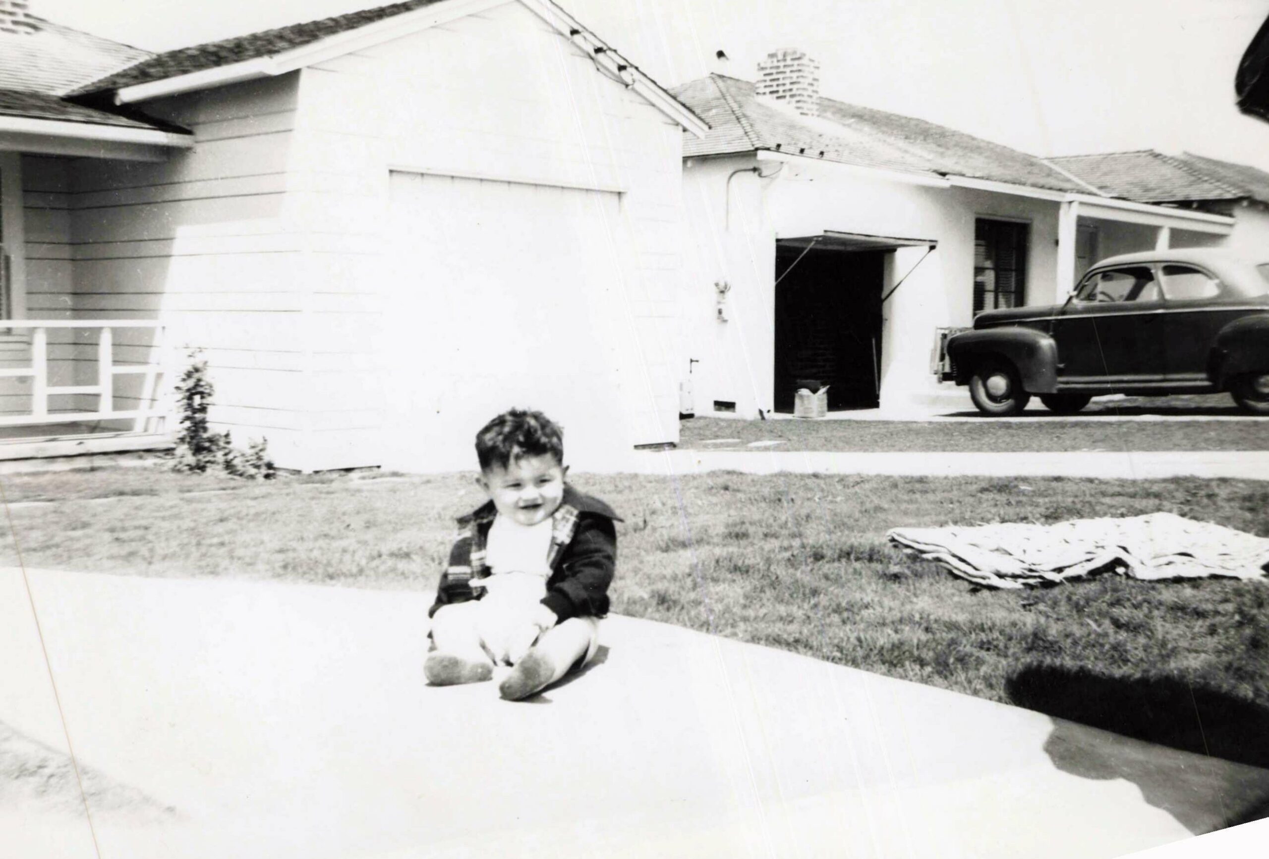 Young child sitting on a concrete step in front of a suburban house, with a vintage car in the driveway to the right in a black-and-white photo.