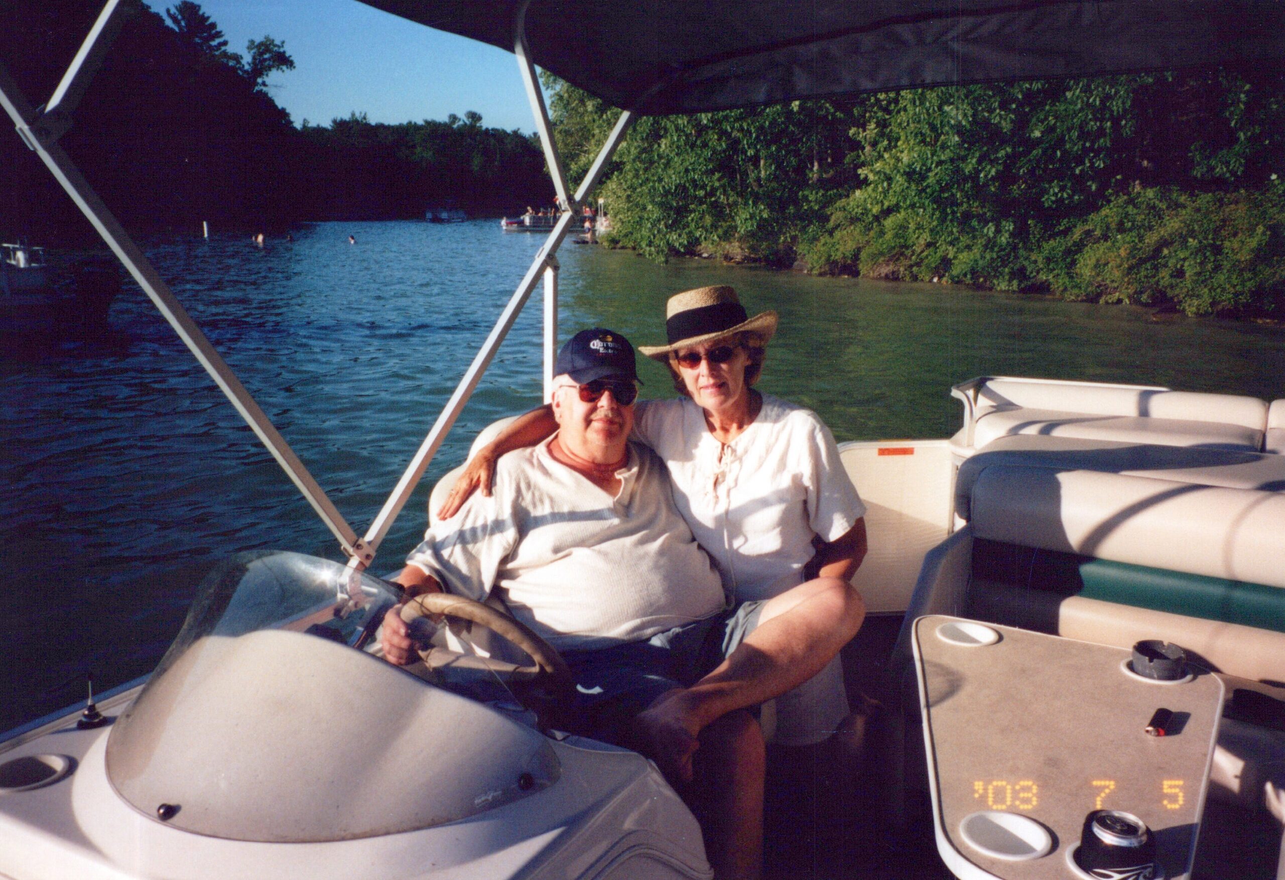 Two men sit close together on a pontoon boat, wearing hats and sunglasses, with a lake and green shoreline in the background.