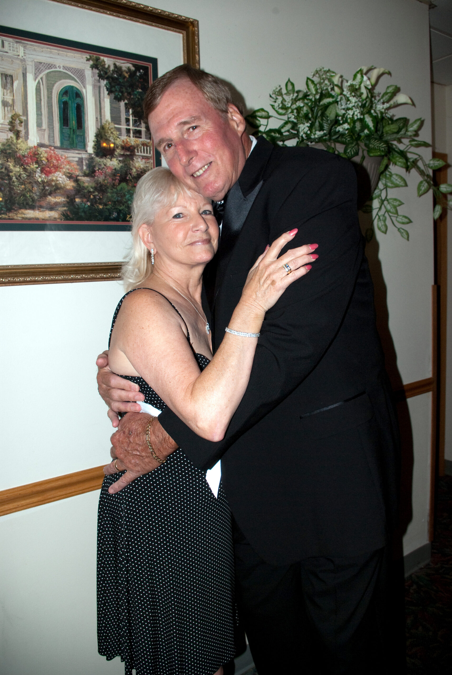 Couple hugging and smiling indoors; woman in a black polka-dot dress, man in a dark suit.