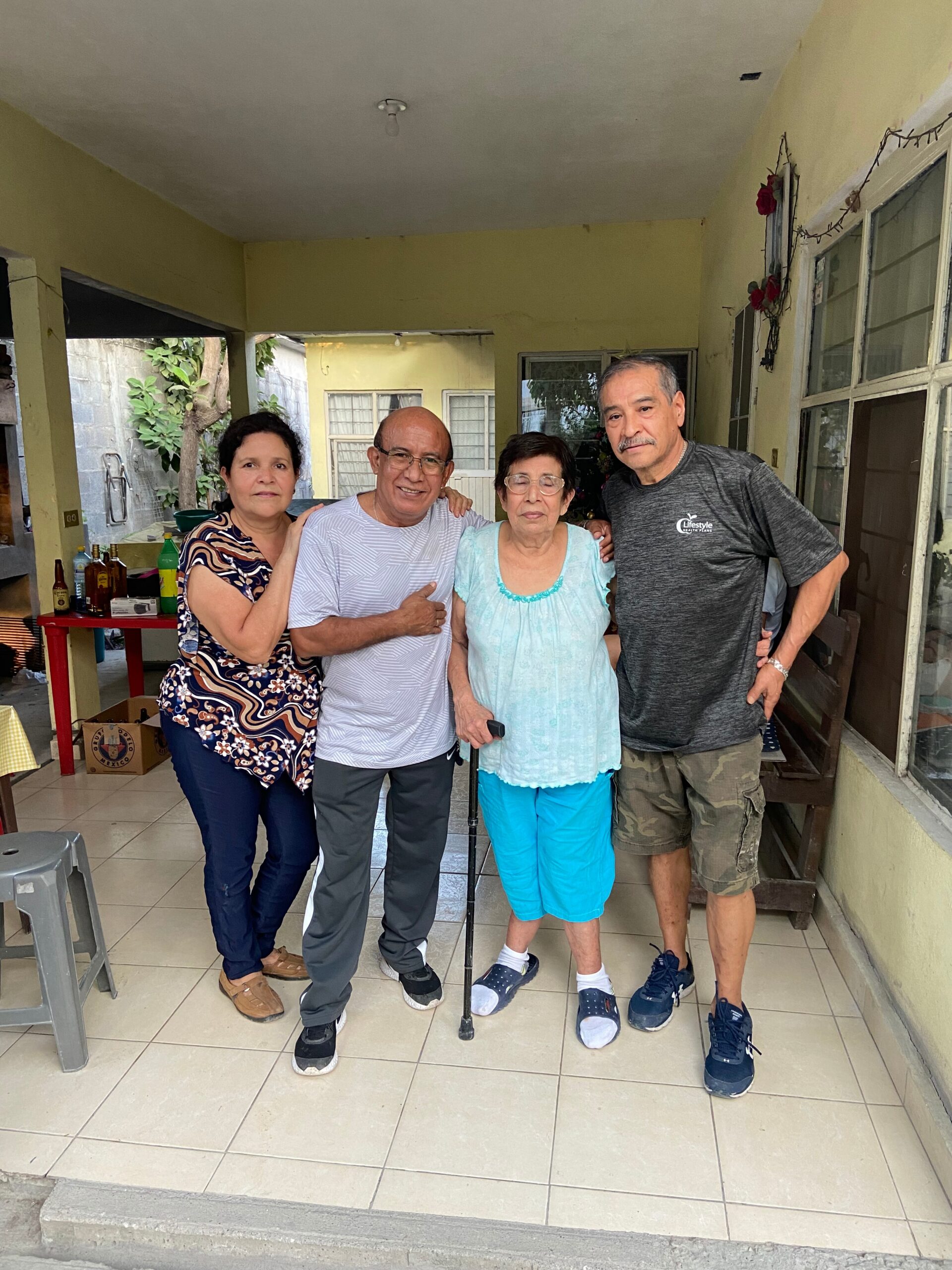 Four adults posing together on a tiled patio outside a yellow house, smiling at the camera.