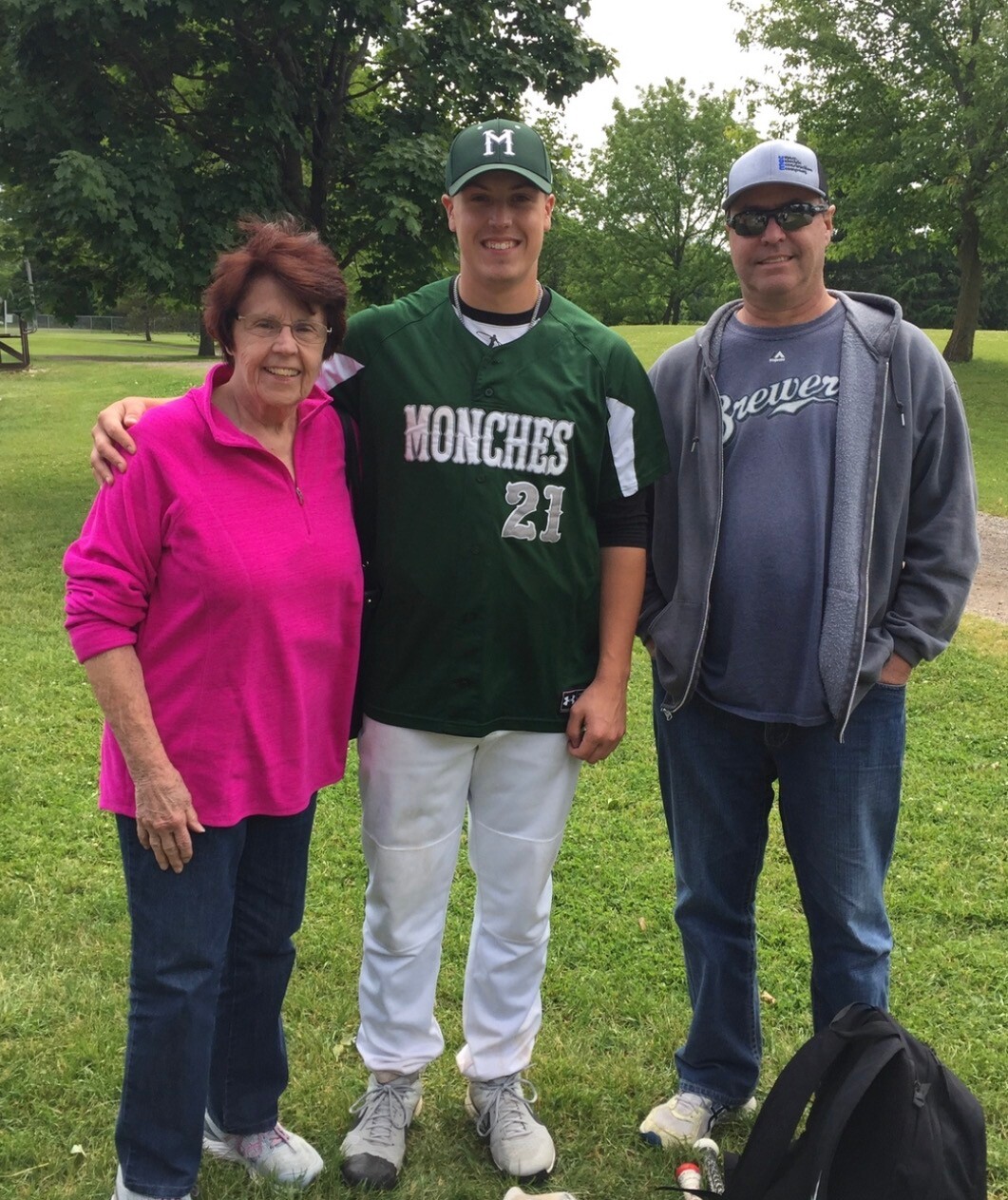 Three people stand together on a grassy park field: an elderly woman in a pink pullover, a smiling baseball player in a green Monches jersey (number 21), and a man in a gray hoodie and sunglasses beside him.