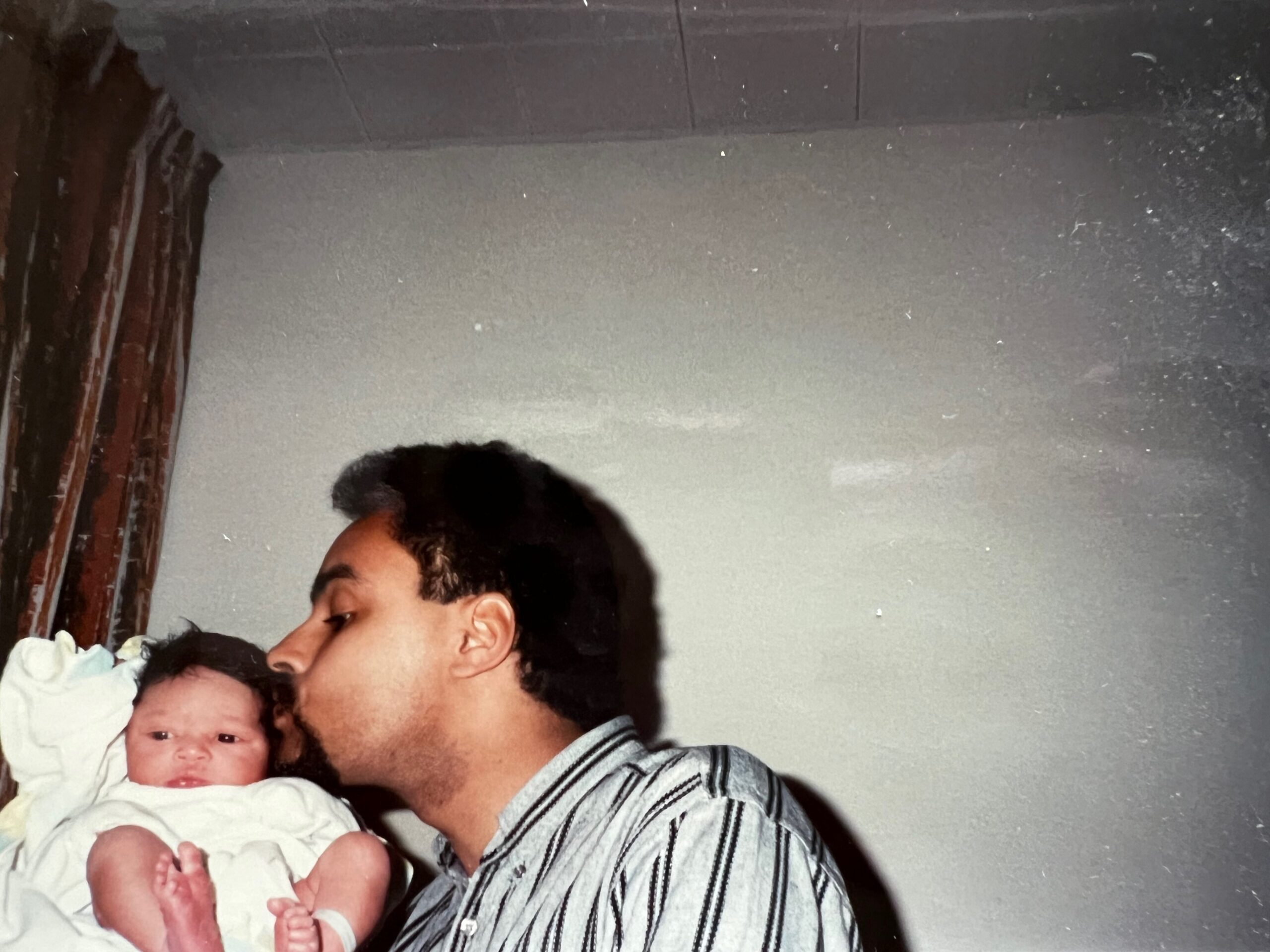 Man in striped shirt holding a newborn baby wrapped in white clothing, indoors against a gray wall with curtains to the left.