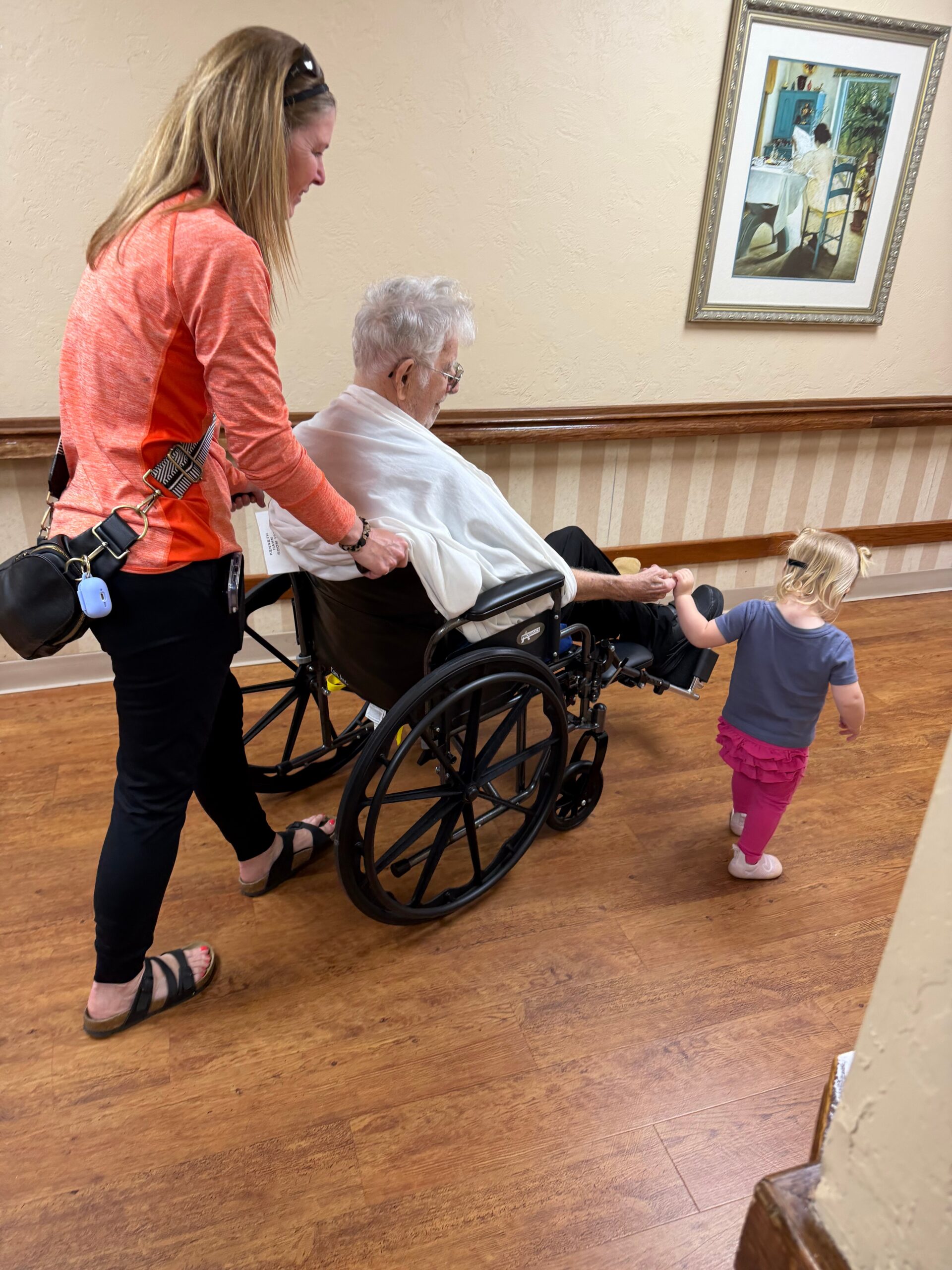 Caregiver in an orange shirt pushes an elderly man in a wheelchair while a young child holds his hand in a hallway, framed painting on the wall behind.